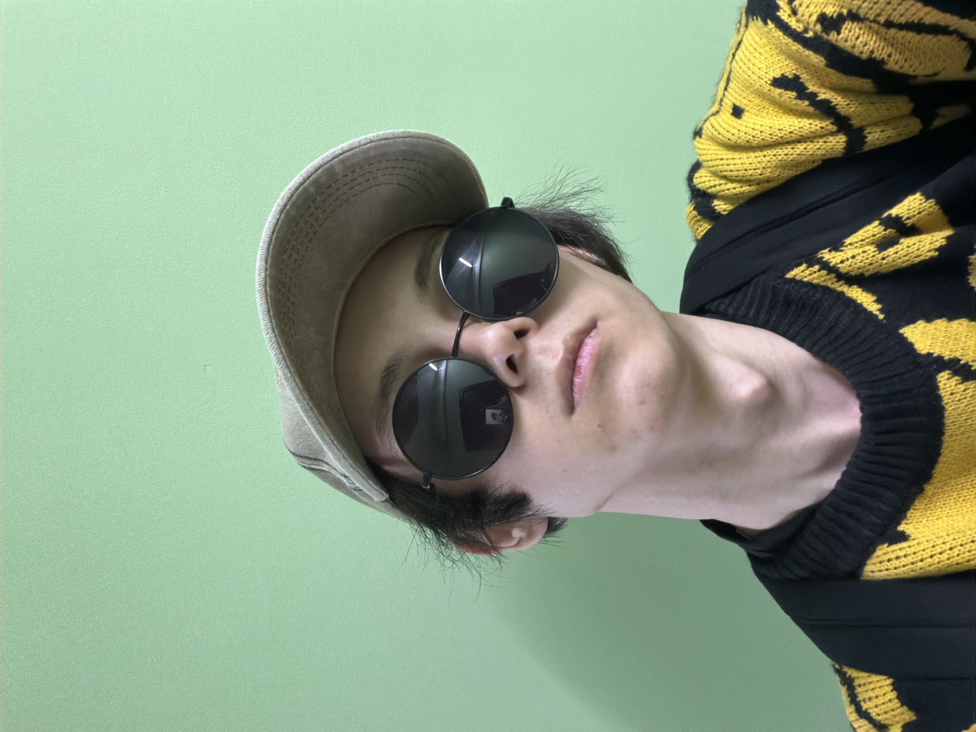 The image shows a close-up, angled shot of a person wearing a baseball cap, sunglasses, and a yellow and black patterned sweater. The person is looking towards the camera, and their face is partially obscured by the accessories. The background is a solid, light green color. The lighting appears to be artificial, suggesting an indoor setting. The person's expression is neutral, and there are no overt emotional cues. The image is well-lit, and the details of the clothing and accessories are clear. There is no visible text in the image.