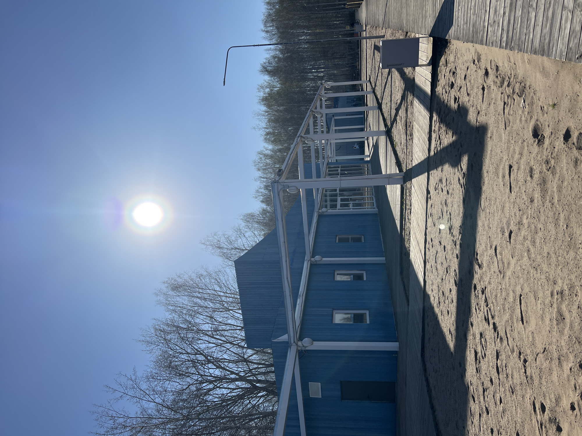 The image shows a building with blue wooden siding in the city of Chaikovsky, Russia. The building has white framing and a few windows. Next to the building is a boardwalk and a sandy area with footprints. Trees are visible in the background. The sun is high in the sky. A light pole is visible next to the building. A small rectangular box is positioned on the boardwalk.