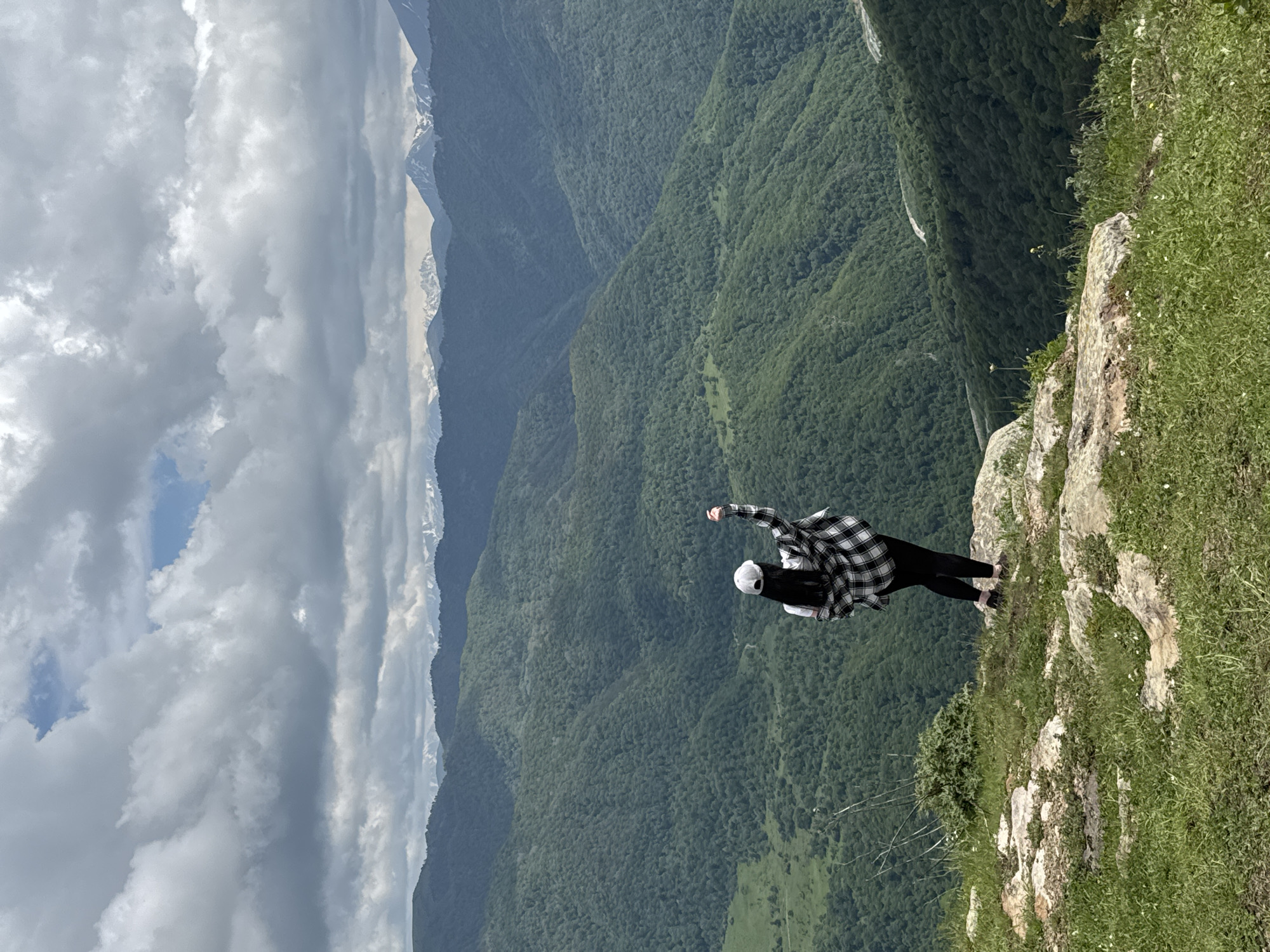 A person stands on the edge of a rocky cliff overlooking a vast, green mountain landscape. The person, viewed from behind, is wearing a white baseball cap, a black and white checkered shirt over a white t-shirt, black pants, and dark shoes. Their right arm is raised, and their fist is clenched. The mountains are covered in dense green forest, with layers of ridges receding into the distance. The sky is cloudy, with patches of blue visible. In the far distance, snow-capped peaks are visible. The foreground consists of grass and rocks. The location is identified as Nizhnyaya Teberda, Russia.