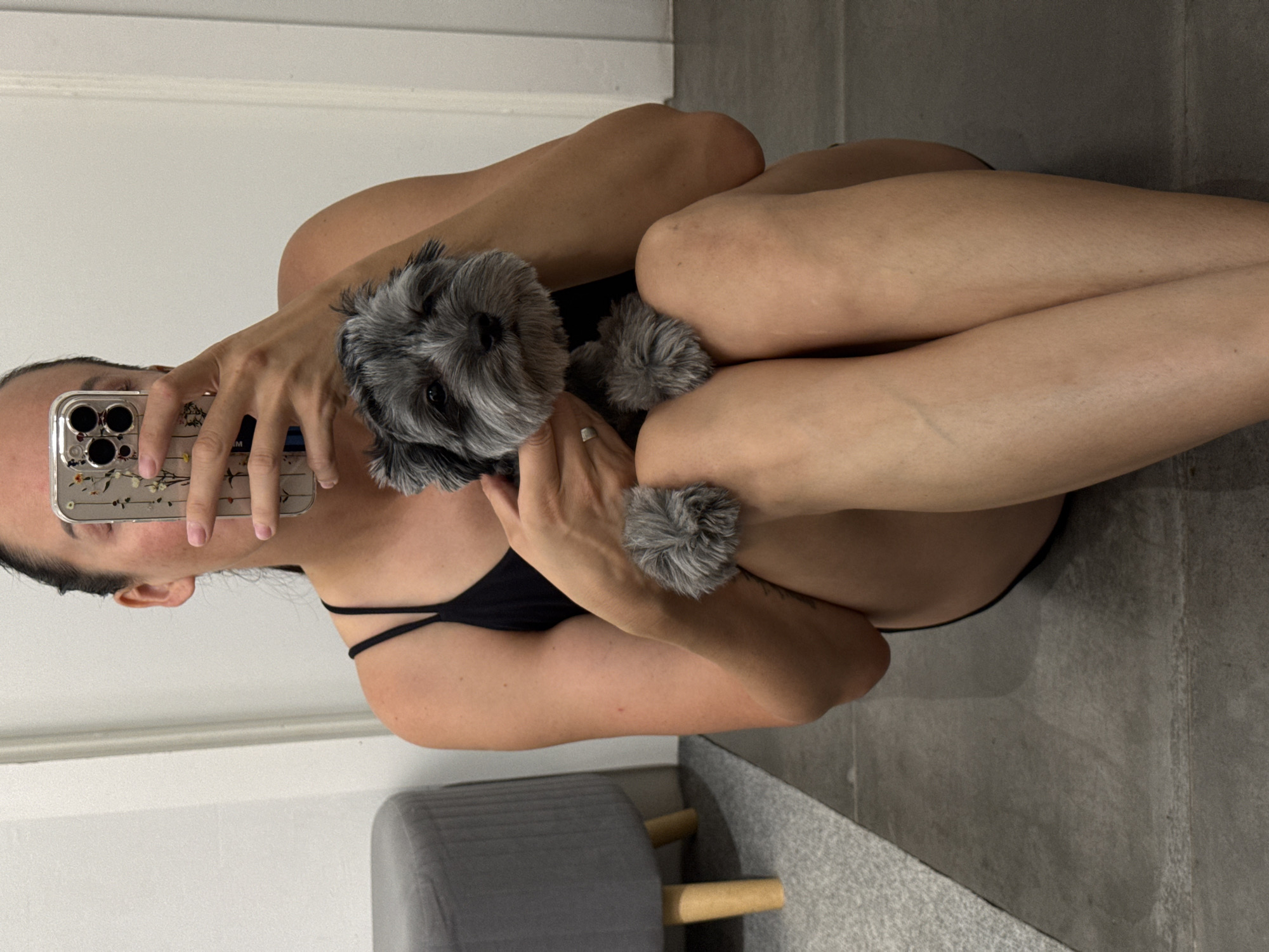 In an indoor setting with gray tiled flooring and white walls, a person is sitting on the floor, holding a small gray dog. The person is wearing a black tank top and is holding a smartphone with a floral case to take a mirror selfie. The dog, which appears to be a Shih Tzu or similar breed, has its paws resting on the person's legs. To the left of the frame, a gray ottoman with wooden legs is visible, along with a gray rug. The scene suggests a casual, home environment. There is no indication of the location being Chaikovsky, Russia.