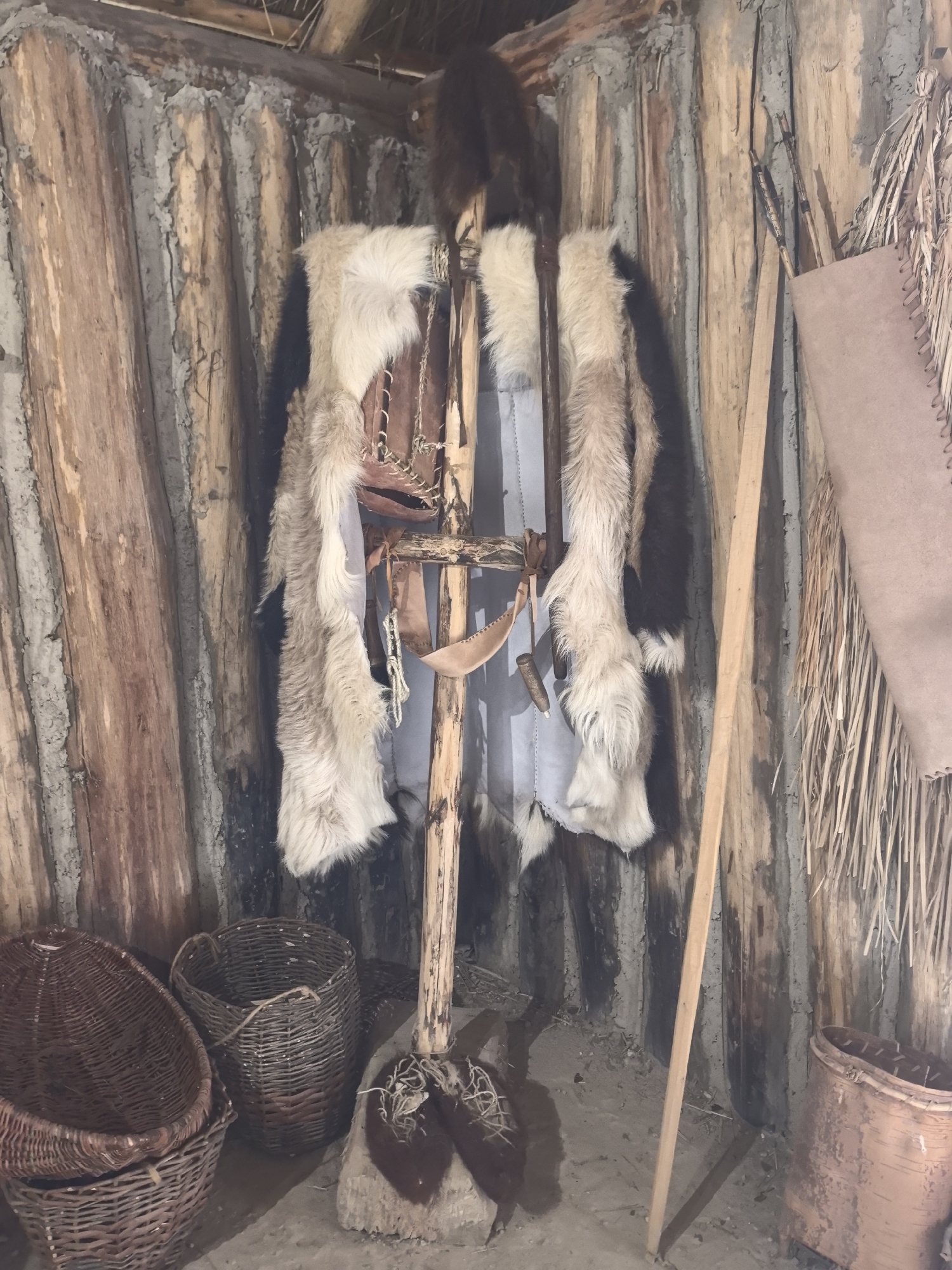 An indoor, full, medium shot shows a display of primitive clothing and artifacts within a structure made of rough-hewn logs and plastered mud. The clothing, consisting of animal furs and what appears to be leather, hangs from a wooden pole standing upright in the center of the frame. A large piece of hide, possibly for a shield or tent, is propped against the wall on the right side, with a bundle of dried reeds or straw to its left. In the foreground, two woven baskets are visible on the left, and a third, larger woven basket or container is on the right. The floor is a sandy, dirt surface. The overall impression is of a historical or archaeological exhibit depicting a dwelling or personal items from an early period.