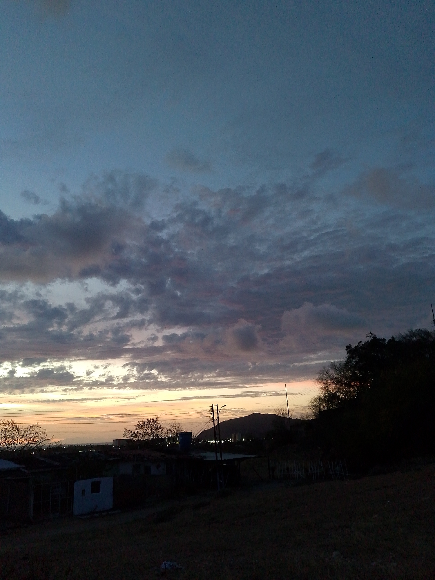 The image depicts a twilight scene in Juan Griego, Venezuela. The sky is filled with layered clouds, colored with shades of blue, grey, and pink from the setting sun. Below the sky, there's a dark landscape featuring silhouetted trees, buildings, and power lines. A distant mountain can be seen. The foreground is dark, suggesting a grassy or undeveloped area. There are no people visible in the image.