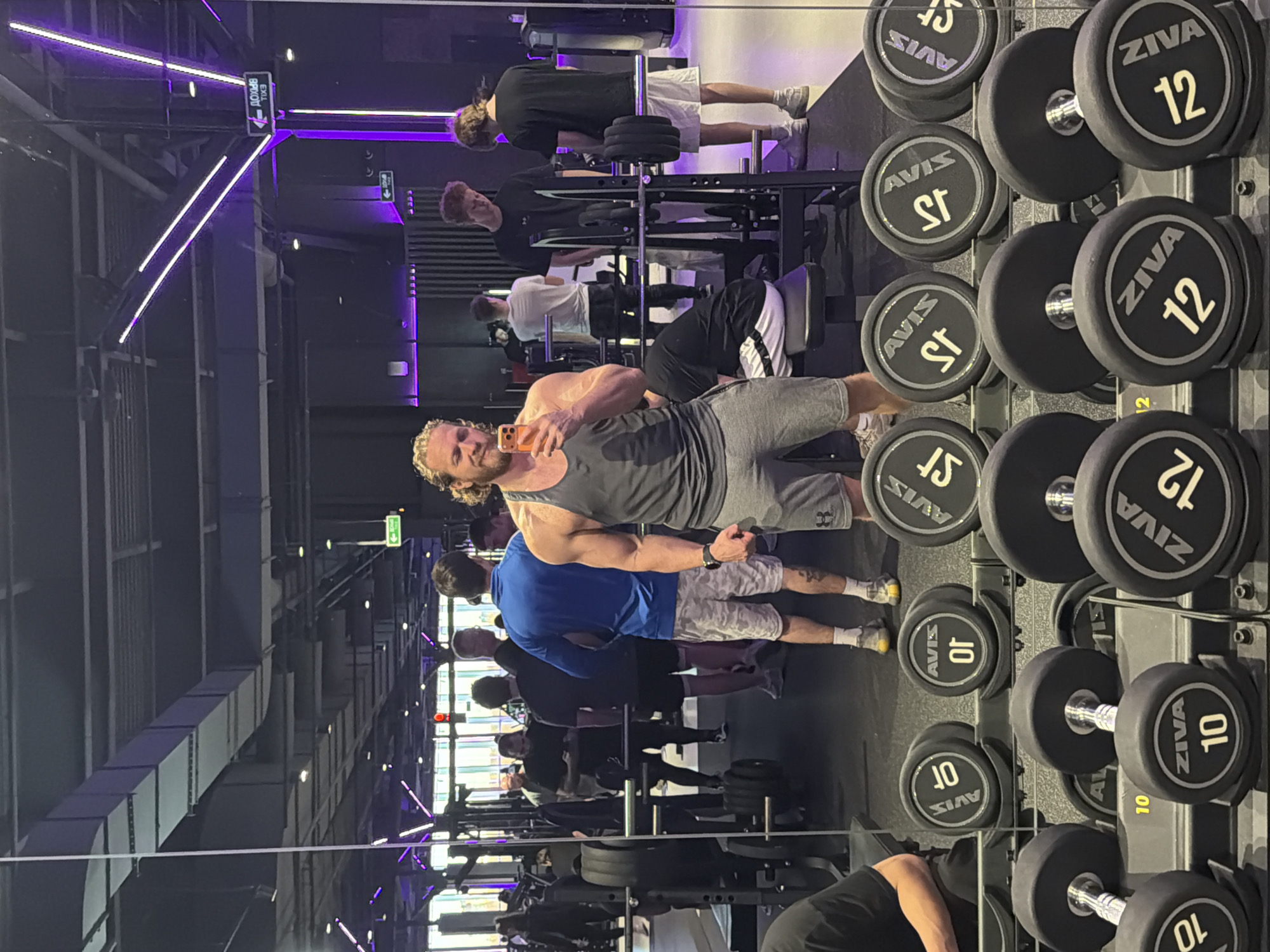 The image depicts an indoor gym scene, likely in Saint Petersburg, Russia, with a man taking a mirror selfie in the foreground. The gym features modern equipment and purple accent lighting on the ceiling, creating a distinct aesthetic.
In the foreground, a fair-haired man with a beard, wearing a grey tank top and shorts, holds a smartphone with an orange case to capture his reflection. He stands beside a rack of dumbbells. The mirror reflects several other gym patrons in the background, engaged in various workout activities, including using weight machines and standing near free weights. One individual in the reflection wears a black t-shirt and white shorts, another a blue t-shirt and camouflage-patterned shorts.
A prominent dumbbell rack is visible on the right, featuring dumbbells from brands like ZIVA and AVIS. Specific weights clearly visible include "ZIVA 12", "ZIVA 10", and "AVIS 10". Other AVIS dumbbells display markings for "15" and "17.5", possibly using local notation.
Above, structural elements, ventilation ducts, and more of the purple strip lighting are visible. A directional sign is clearly seen with Cyrillic text "ВХОД" (Entrance) and "EXIT" below it, accompanied by arrows, confirming the Russian-speaking location. The scene portrays an active fitness environment.