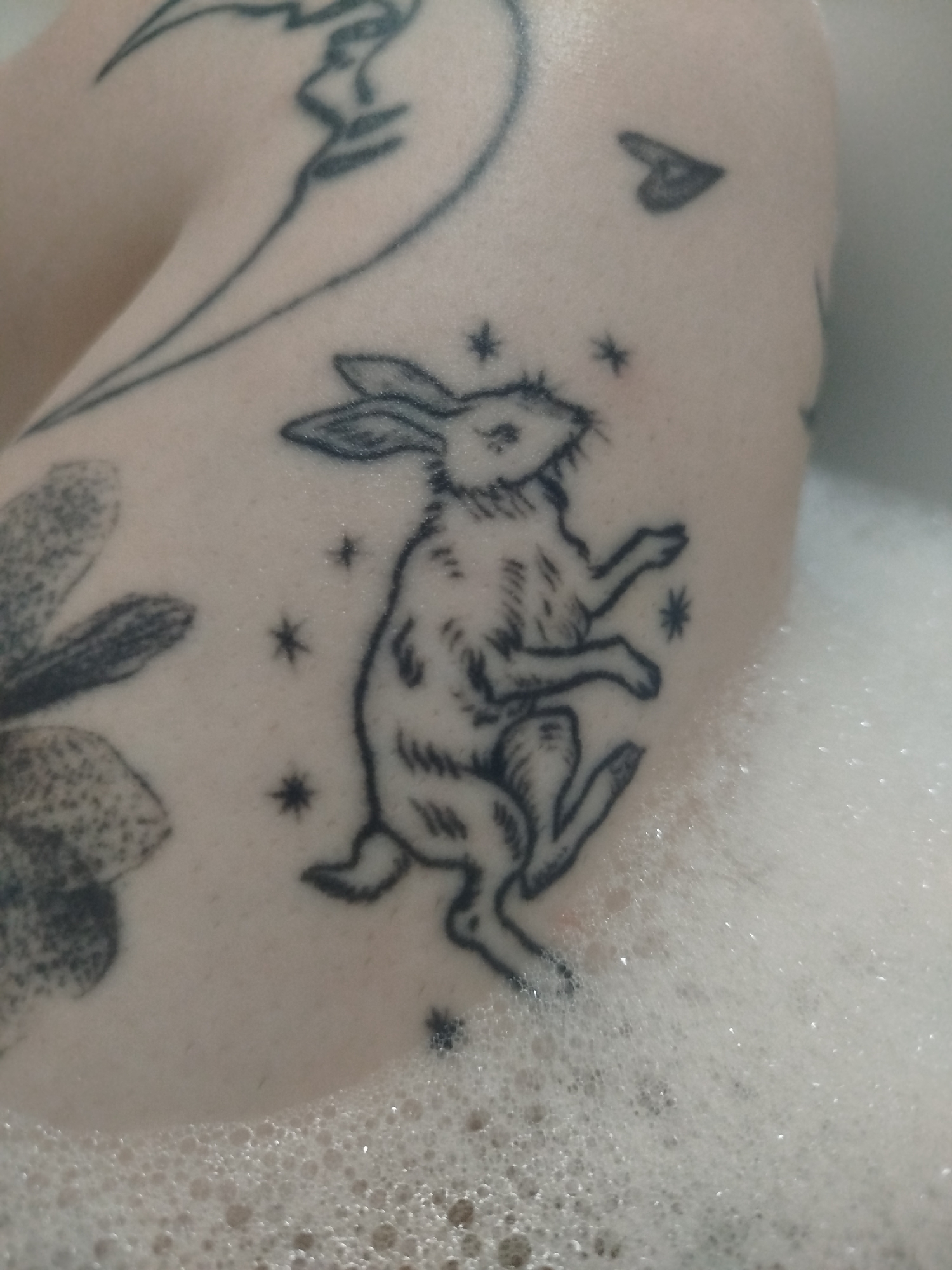 The image shows a person's arm in a bathtub filled with bubbly water. The arm has several tattoos, including a rabbit standing on its hind legs surrounded by stars, a flower, a heart, and an abstract design. The tattoos are black ink. The environment is a white bathtub with foamy water. The location is not specified, and thus is unknown.