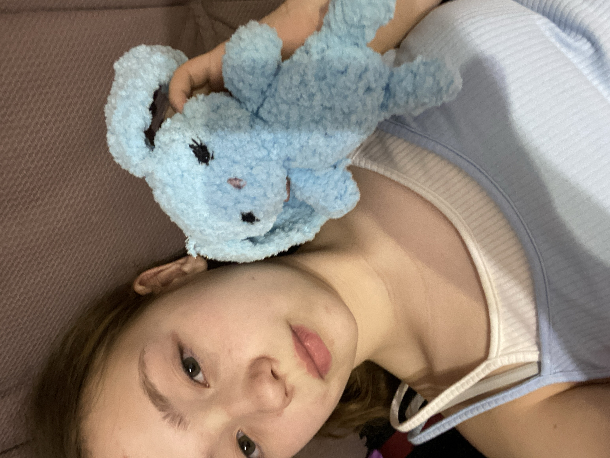 A close-up, angled shot shows a young person with light skin lying down, possibly on a couch based on the neutral-toned textured fabric visible in the background. The person has brown hair and green eyes, and their face is turned towards the camera. Atop their head is a small, light blue stuffed animal with black eyes and a stitched nose. The person is wearing a light blue and white tank top. The location and city are unknown.