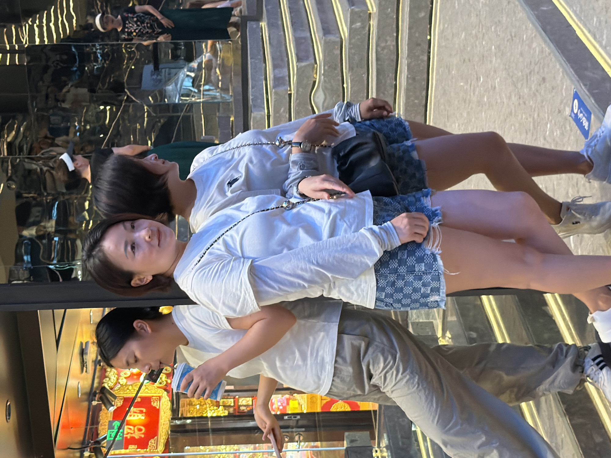 The image shows a group of people in an urban environment, possibly in Yuzhong District, China. In the foreground, two young women are standing. The woman on the left is wearing a white t-shirt, grey sweatpants, and athletic shoes, and is holding a blue and white cup while looking to her left. The woman on the right is wearing a white long-sleeved shirt, blue denim shorts, and white sneakers, and is looking forward, with a black crossbody bag. In the background, reflections in a mirrored surface reveal other individuals, including a woman in a white visor and a dark top, another woman in a white cap and a patterned top with a dark skirt, and a third person in a dark green garment. The setting appears to be a commercial area with a brightly lit display behind the woman on the left, and a set of illuminated stairs in the background.
