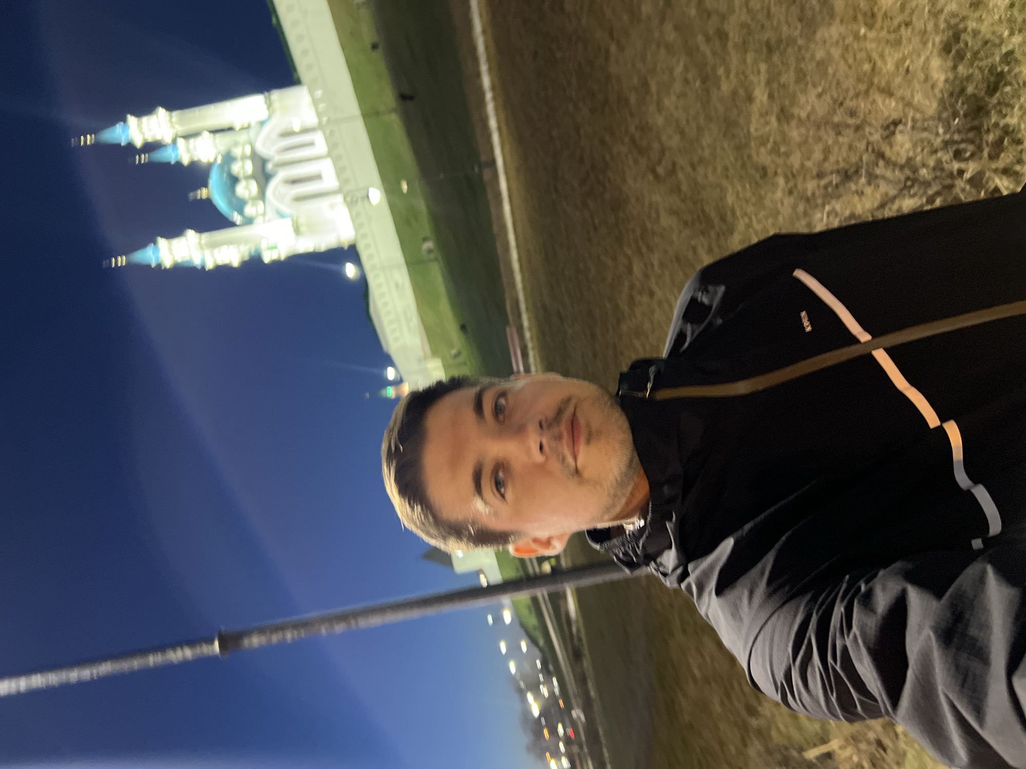 The image shows a man posing for a selfie at night. Behind him, illuminated, is the Qolşärif Mosque in the Kazan Kremlin, Kazan, Russia. The surrounding area includes grass and streetlights. The sky is a deep blue. The man is wearing a black jacket.