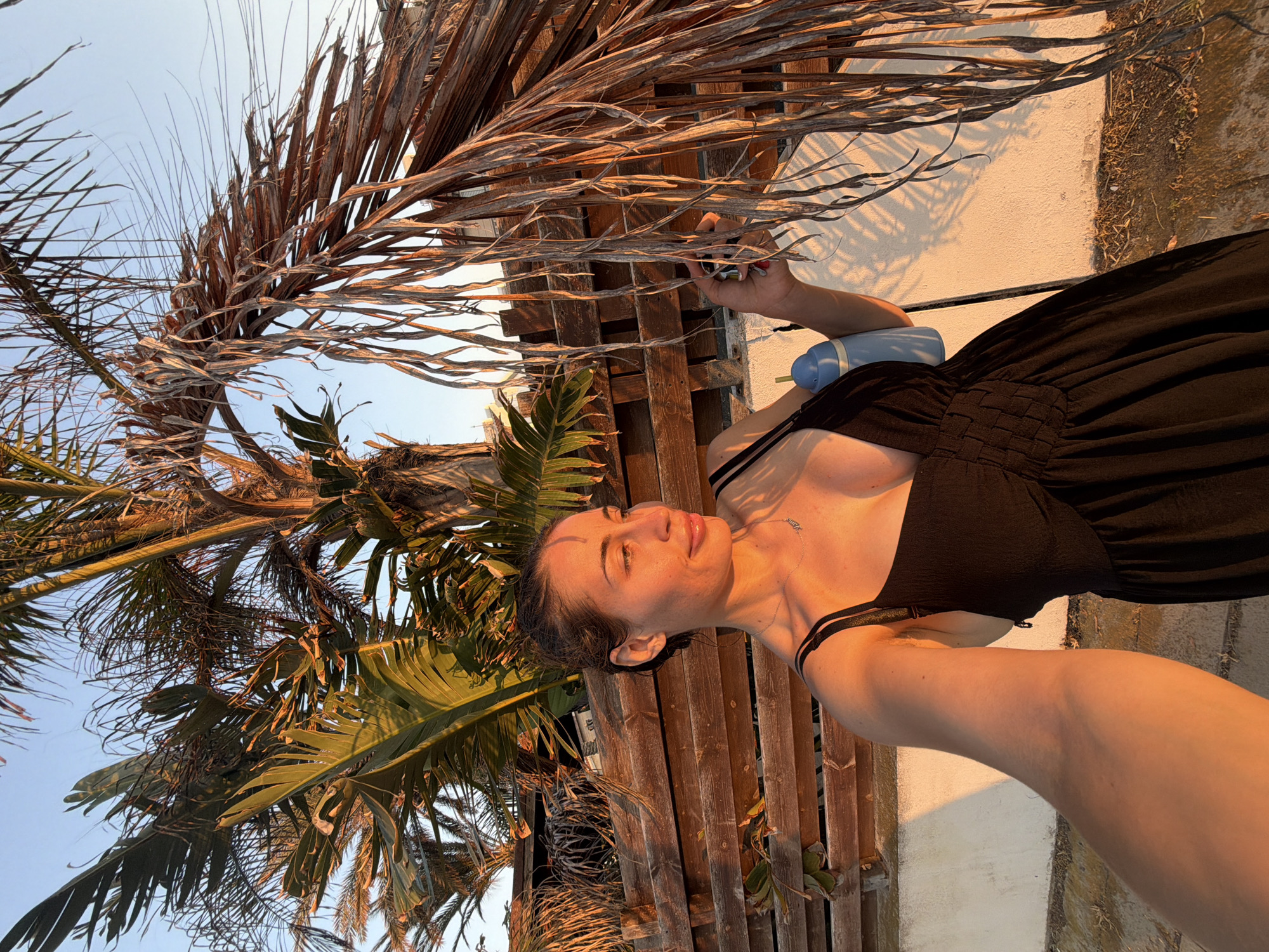 A woman in a black dress stands outdoors in Meneou, Cyprus, under a palm tree. The sun is setting, casting a warm glow on the scene. The woman is holding a light blue object in her right hand. A wooden fence is visible behind her, and a white wall is to her right. The ground is covered in dry leaves and dirt. The sky is clear and blue.