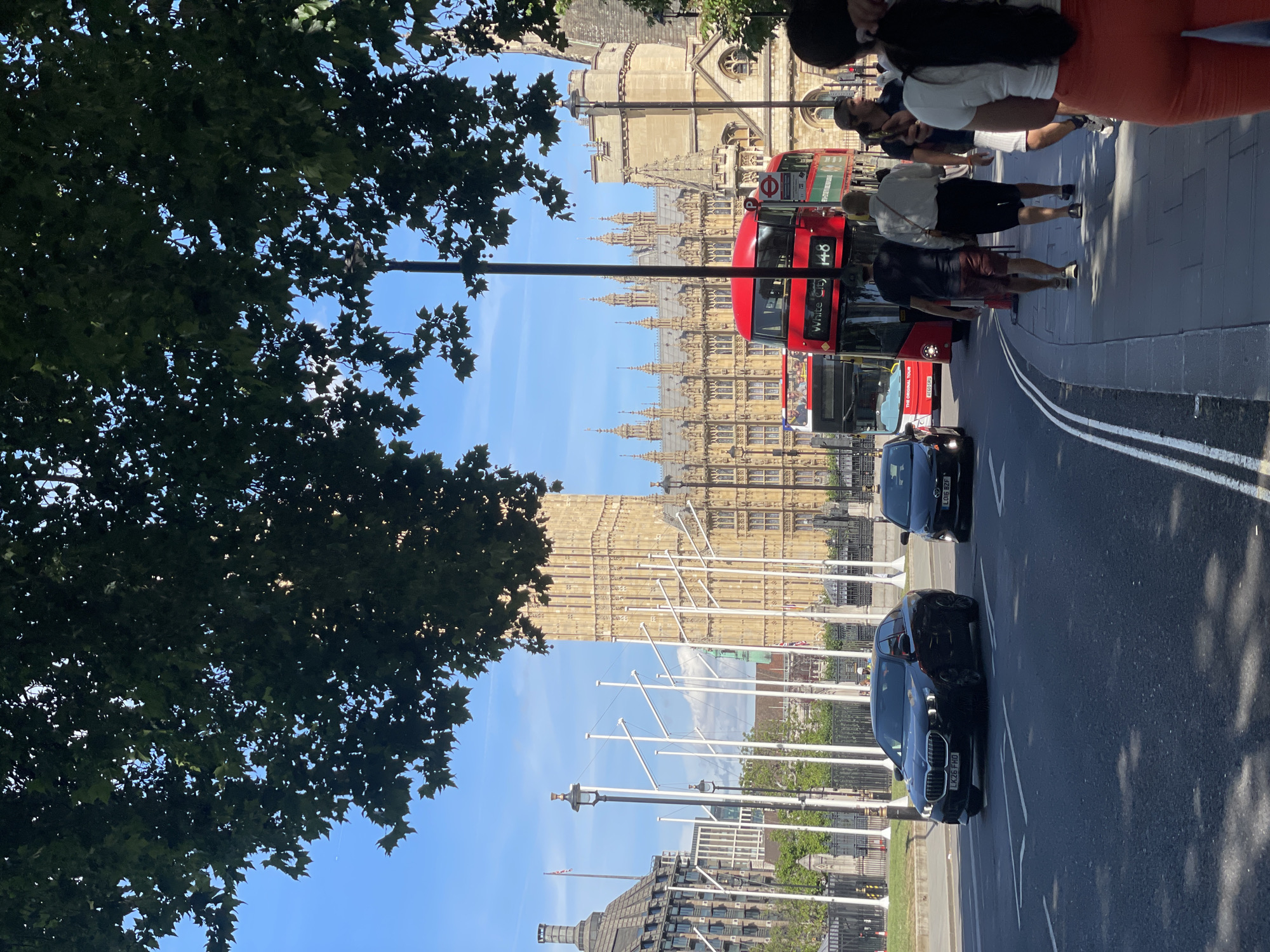 In the city of London, United Kingdom, a street scene unfolds with the iconic Palace of Westminster and Elizabeth Tower as a backdrop. The image captures a sunny day with a clear blue sky, partially obscured by the lush green foliage of trees in the foreground. A black BMW sedan is driving on the road, followed by a red double-decker bus. A red open-top tour bus is also visible behind the double-decker. Several pedestrians are walking on the sidewalk, including a woman in an orange outfit, a man in a white shirt and black shorts, and another man in a black t-shirt and brown shorts. Streetlights and flagpoles line the sidewalk. The architecture of the Palace of Westminster, with its numerous spires and Gothic style, dominates the background. The general atmosphere suggests a bustling tourist area.