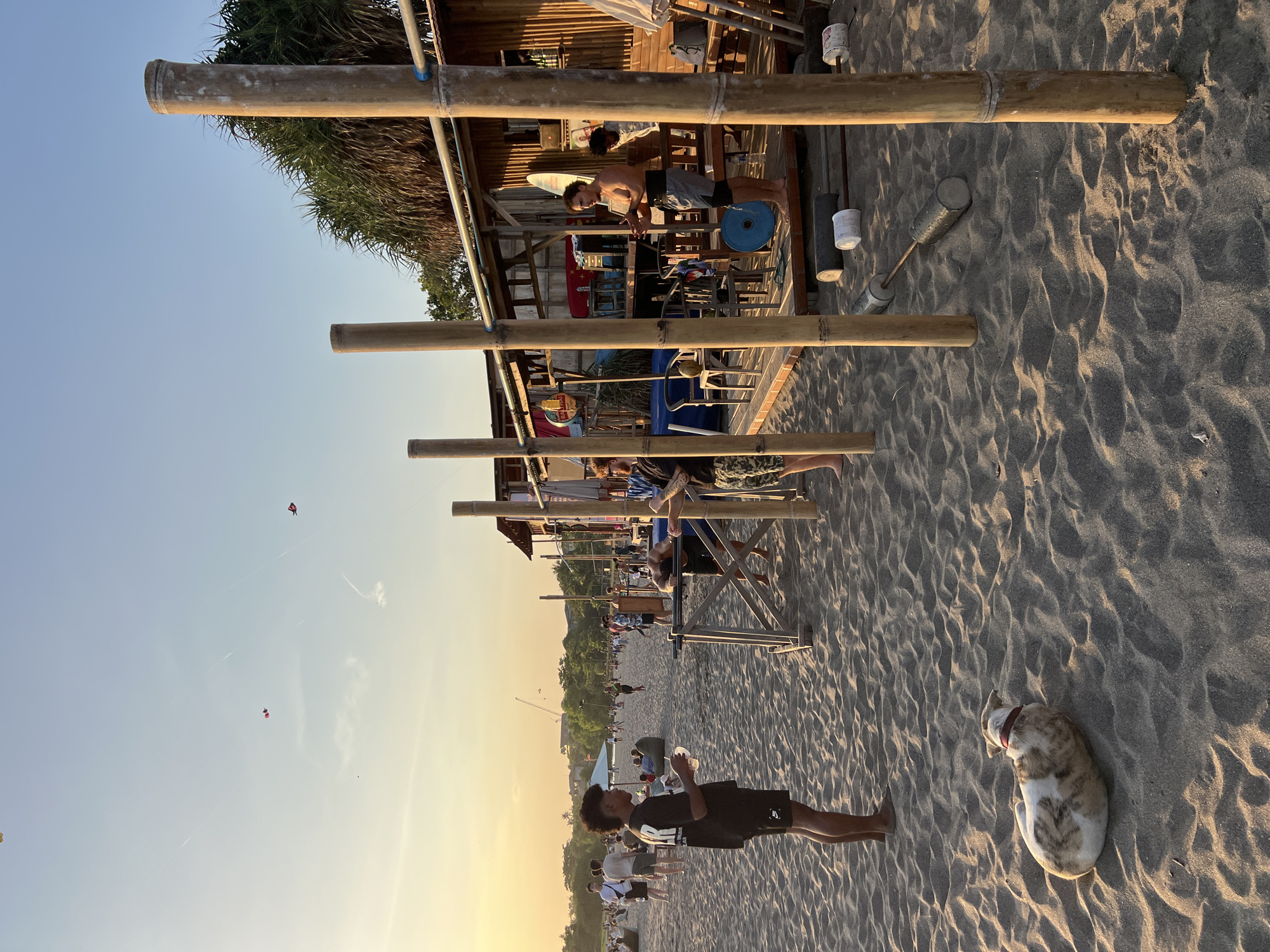 This image depicts a beach scene at sunset in North Kuta, Indonesia. The foreground shows a sandy beach with a dog lying down. Several people are visible in the midground and background, some standing and interacting, while others are engaged in various activities. A bamboo structure, possibly a bar or food stall, is present on the right side of the frame, with a few individuals near it. There is a clear sky with kites visible in the distance. The lighting suggests it is late afternoon or early evening. The overall atmosphere appears relaxed and social.