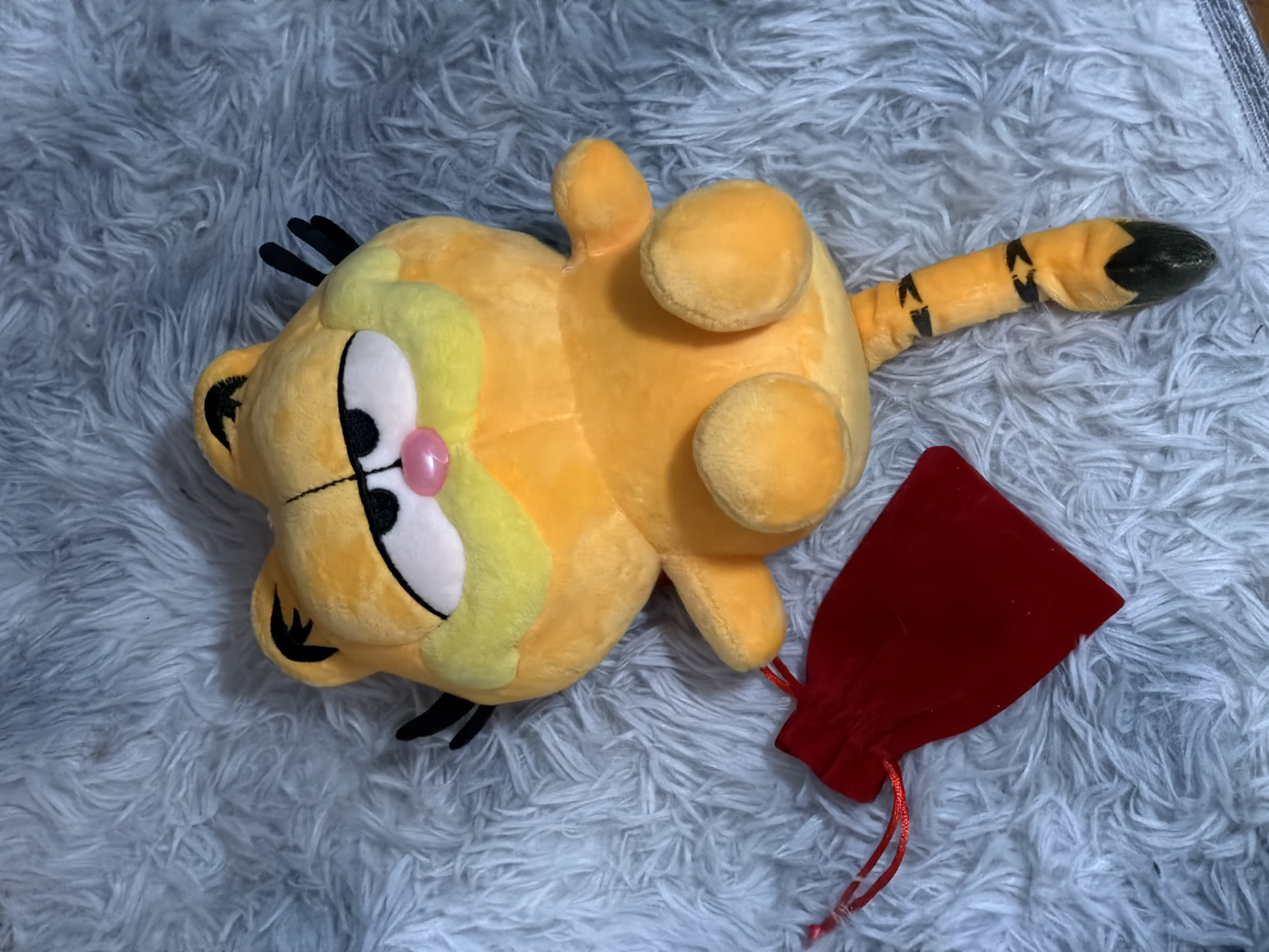 A Garfield plush toy is lying on a fluffy, white rug. The toy is orange and yellow with black stripes on its tail. To the left of the Garfield toy is a small, red velvet drawstring pouch. The background is a blurry, white, fluffy rug. There is no indication of Moscow, Russia in the image.