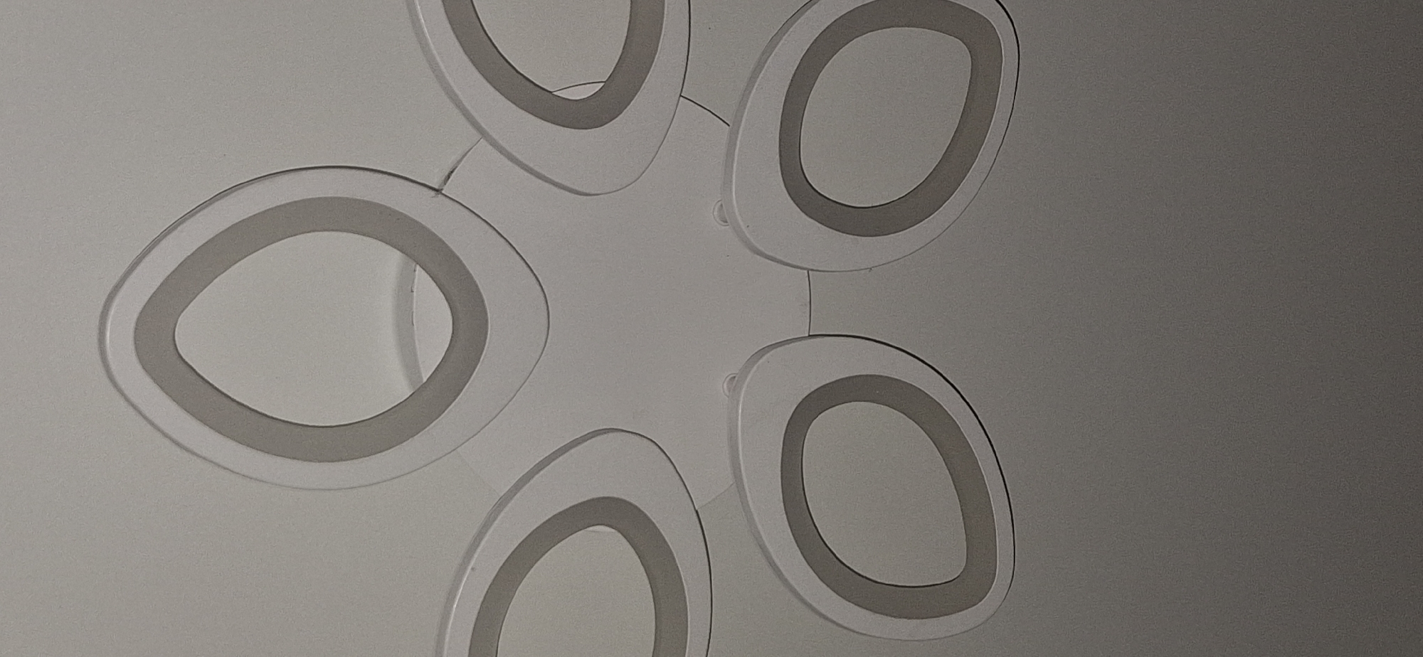 This is a close-up, overhead, indoor photograph of a modern, white ceiling light fixture. The fixture has a central circular base from which five oval-shaped light elements extend. Each light element is a flat, white, oval-shaped piece with a recessed inner oval outlined in a light grey color. The light elements are positioned radially around the central base, with three in the upper half and two in the lower half of the frame, suggesting a symmetrical arrangement. The background is a plain white ceiling. There are no people, animals, or any other objects visible in the image. The image does not provide any information about the city of Bila Tserkva, Ukraine.