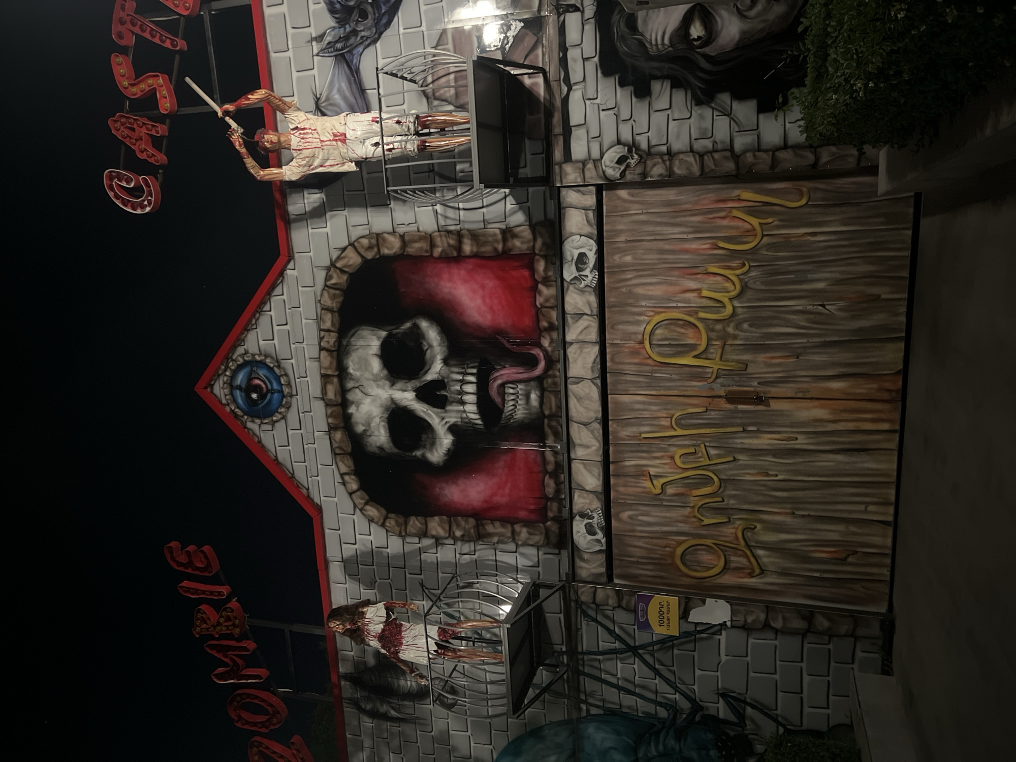 The image displays the illuminated facade of a "Zombie Castle" attraction at night, located in Yerevan, Armenia, as evidenced by the Armenian script "Զոմբի Քասթլ" (Zombie Castle) on the main doors and a price sign indicating "1000դր" (1000 Armenian dram) for "1 անձի համար" (one person).
The structure is designed to mimic a multi-level, grey brick castle or mansion, with a red-trimmed roofline. Large, illuminated red letters spelling "ZOMBIE CASTLE" are prominently displayed at the top against a dark night sky. A central dormer on the roof features an illuminated blue eye graphic.
Two blood-splattered, zombie-like mannequins are mounted on elevated metal platforms. On the left, a female mannequin in a torn white dress stands on a circular platform. On the right, a male mannequin in torn white clothing, holding a white blunt object above its head, stands on a rectangular platform.
Below the roof, a large central mural depicts a skull with a prominent tongue, set within a stone archway against a dark red background. Smaller skull designs are painted on the facade sections below this main mural. A partial depiction of a grotesque face with dark hair is visible on the far right side of the facade.
The main entrance consists of large, double wooden-textured doors, upon which "Զոմբի Քասթլ" is painted in stylized yellow script. To the left of the entrance, a small purple and yellow sign displays the admission price. The lower sections of the facade are flanked by green foliage, and the foreground consists of a dark paved area.