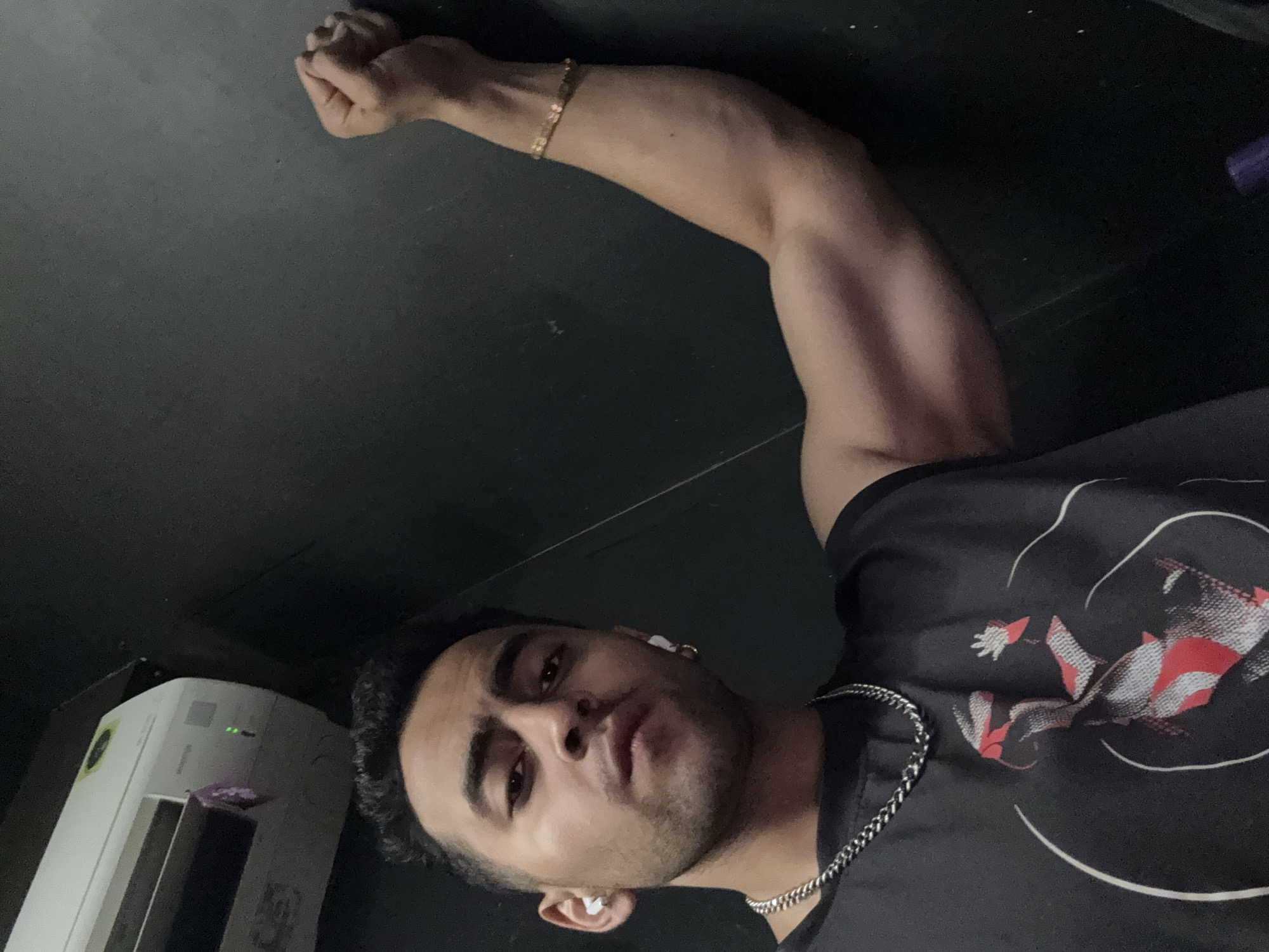 A medium shot depicts a man in what appears to be an indoor setting. The man has dark hair, light brown skin, and is wearing a black T-shirt with a white and red graphic design. He wears silver chain, and ear buds. The man is posing with his right arm raised, flexing his bicep. The background is dark and indistinct, possibly a wall or ceiling. To the left of the man, there's an air purifier. Based on the visible elements, there's no indication of Kolkata or India.