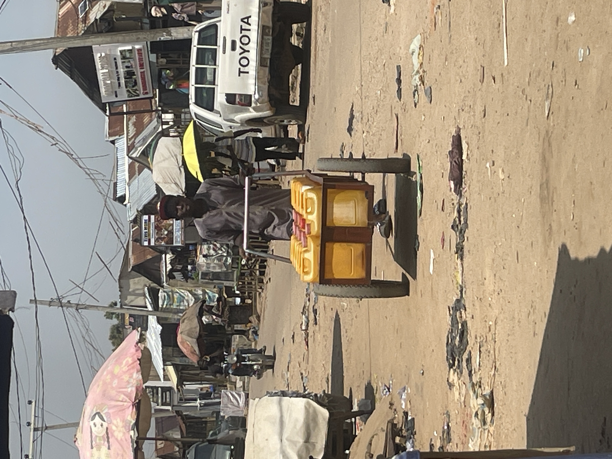 In Abuja, Nigeria, a man pushes a cart laden with yellow containers through a dusty, unpaved street. The cart appears to be used for transporting liquids, possibly water or other commodities. The man, dressed in a dark shirt and trousers, is the central figure in the foreground. The street is lined with various structures, including stalls and buildings with corrugated metal roofs, suggesting a market or a bustling commercial area. A white Toyota pickup truck is parked on the right side of the street. In the background, more stalls and buildings are visible, along with utility poles and overhead wires, indicating an urban or semi-urban setting. The ground is littered with debris and scattered items, adding to the impression of a busy, working environment. Several umbrellas, both open and closed, are scattered throughout the scene, providing shade. A motorcycle is also visible in the middle ground, suggesting ongoing activity and transportation.