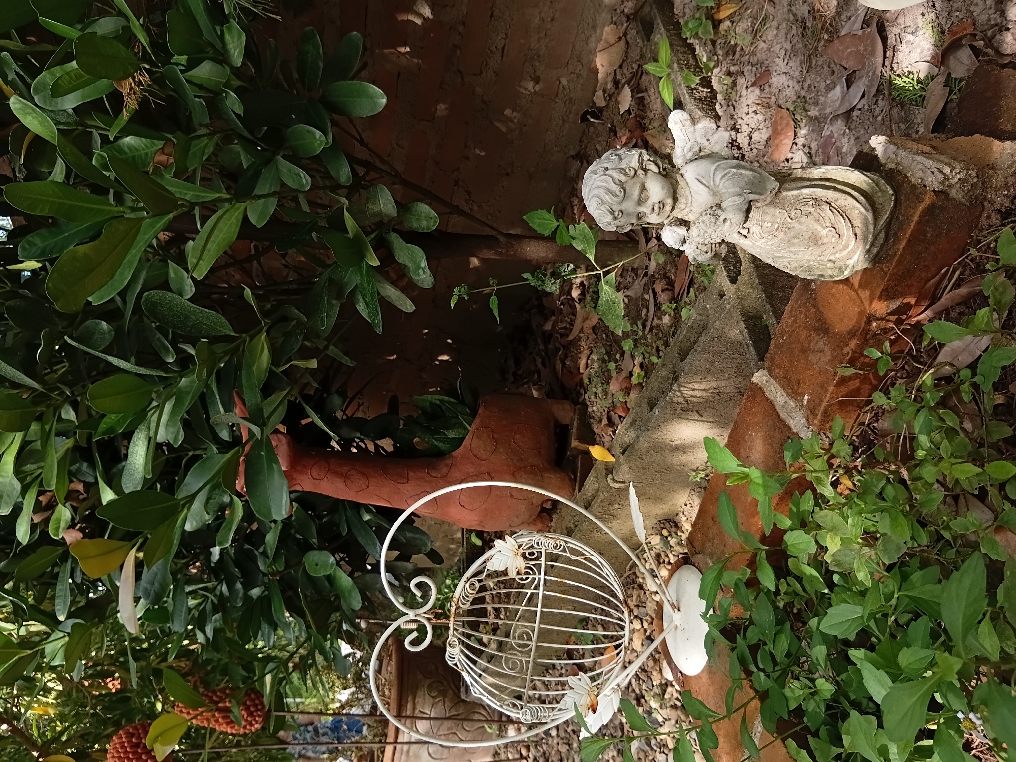 In the city of Ban Tong, Thailand, a garden scene unfolds with various decorative elements. A white metal birdcage, shaped like a heart, stands on a gravel path, adorned with two white floral accents. Behind it, a terracotta giraffe planter is positioned near a lush green tree. To the right of the birdcage, a small cherub statue, made of stone, is placed on a brick. The ground is covered with a mixture of dirt, fallen leaves, and small stones, with green plants growing around the brick and the cherub. In the background, more greenery and a brick wall suggest a private outdoor space.