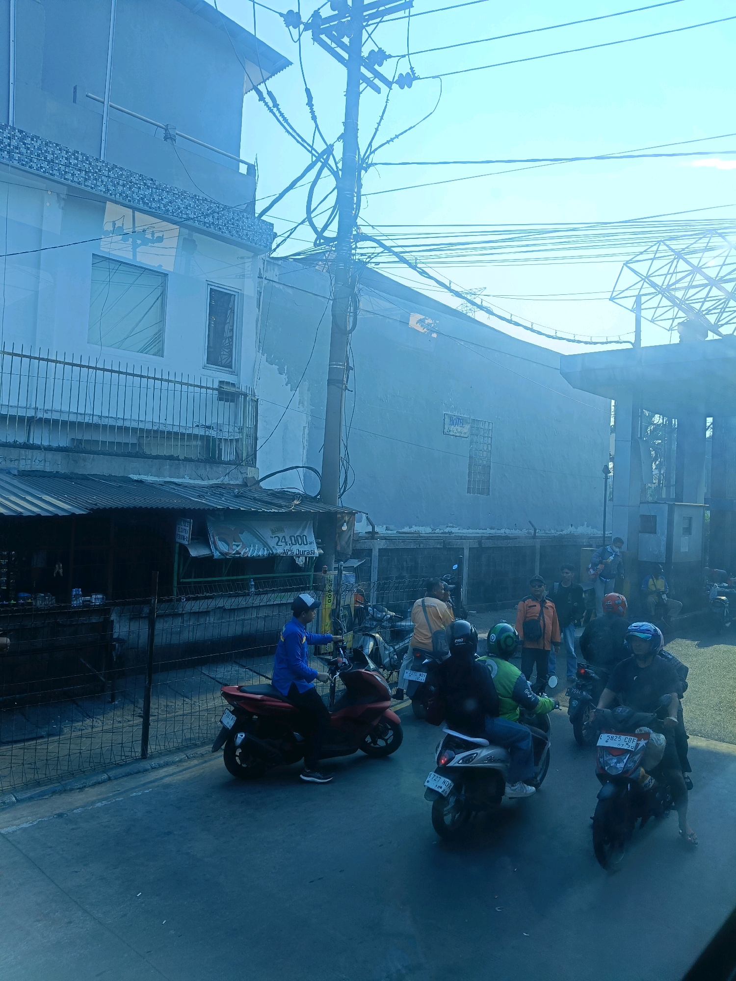 In the city of Kedungrejo, Indonesia, a street scene unfolds under a bright sky. A blue-uniformed individual operates a red scooter, moving along a paved road. Several other motorbikes, some with riders, are visible, indicating moderate traffic. A building with white walls and a corrugated metal roof is situated to the left, with a metal fence in front. Various electrical wires crisscross the sky above the street. In the background, a large concrete structure, possibly part of an overpass or elevated road, dominates the right side of the frame. The overall impression is that of a bustling urban environment with typical street activity and infrastructure.