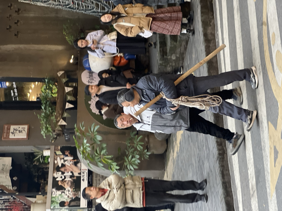 The scene depicts a pedestrian area in the city of Liangjiang, China. In the foreground, two men are walking from left to right across a yellow and white striped pedestrian crossing. The man on the left wears a grey and white jacket, black pants, and sneakers, and has a lit cigarette in his mouth, facing the man to his right. The man on the right wears a dark blue jacket, grey pants, sneakers, and grey earmuffs. He carries a long, light-colored wooden pole across his right shoulder, from which a coil of beige rope hangs.
To the left, another man stands with his arms crossed, wearing a beige puffer jacket, red undershirt, dark pants, and dark shoes, observing the scene to his right. In the background, to the right, three individuals are visible on a slightly elevated pavement. A young woman in a light purple jacket and black skirt is looking at a smartphone. Beside her, a young girl in a brown puffer jacket, plaid skirt, and dark shoes is looking towards the left foreground. Behind these individuals and further back on the right, a large structure decorated with string lights, resembling a Christmas tree, is partially visible.
The background features several storefronts. One building on the left displays a large poster with multiple individuals and Chinese characters. Another building facade on the right has potted plants, a "WELCOME" sign with Chinese characters, and small banner flags. A large, light-colored character statue with a blue element is visible behind the walking men, near other partially visible individuals. The ground varies between patterned pavement and the marked pedestrian crossing.
