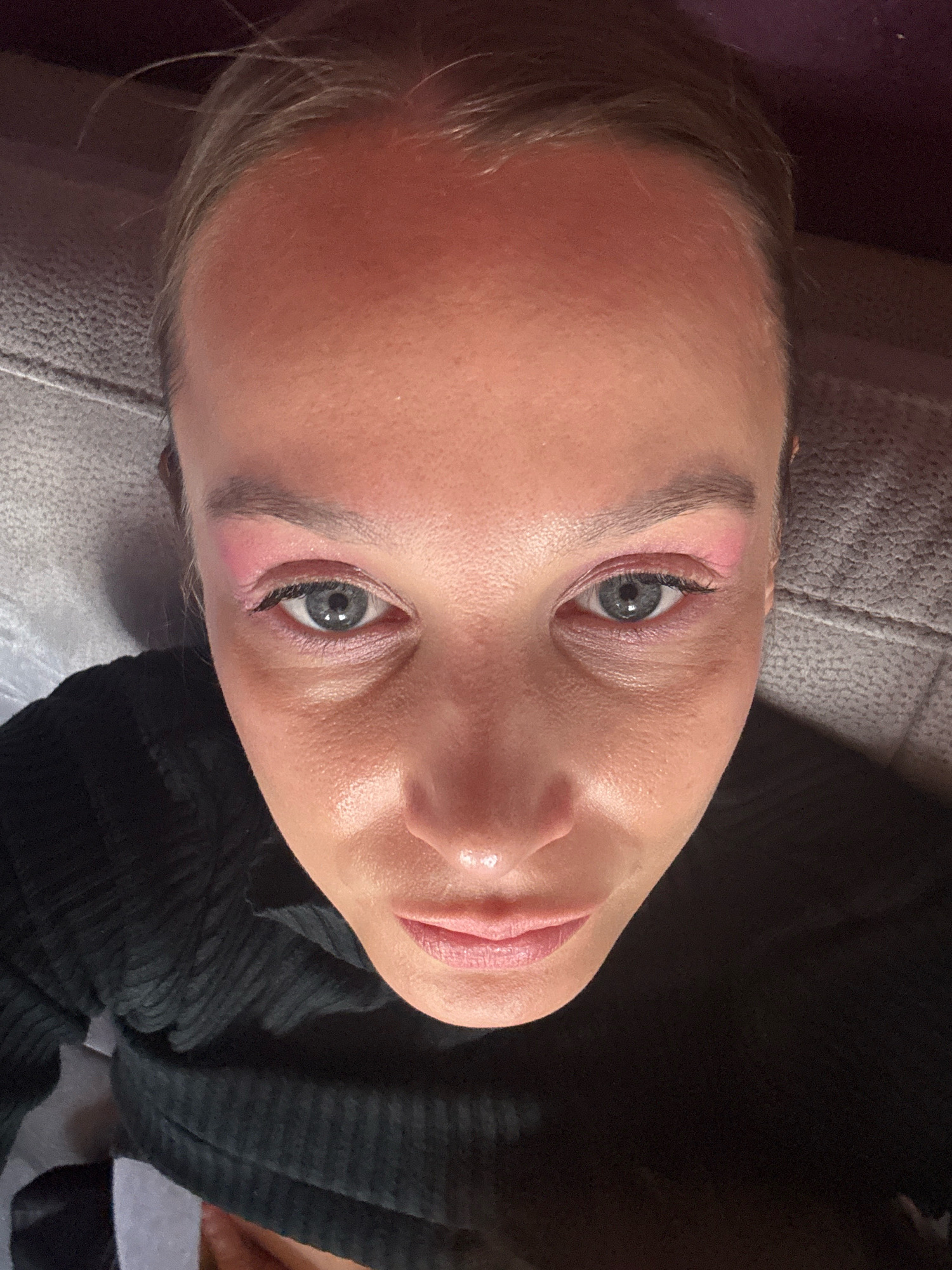A close-up, high-angle shot shows a person's face from the forehead down to the mouth. The person has fair skin with visible pores and some sheen, suggesting oiliness or makeup. Their eyes are open and looking upwards, with gray irises. There is pink eyeshadow applied to the eyelids and extending slightly towards the inner corner of the eyes. Eyeliner is visible on the upper lash line, and lashes appear to be made up. The eyebrows are thin and light brown. The person is wearing a black, ribbed garment that covers their neck and chest. The background appears to be a textured surface, possibly a couch or upholstered furniture, in a muted gray tone, and a darker background on the right. There are no explicit indicators of location in Überherrn, Germany within the image itself.