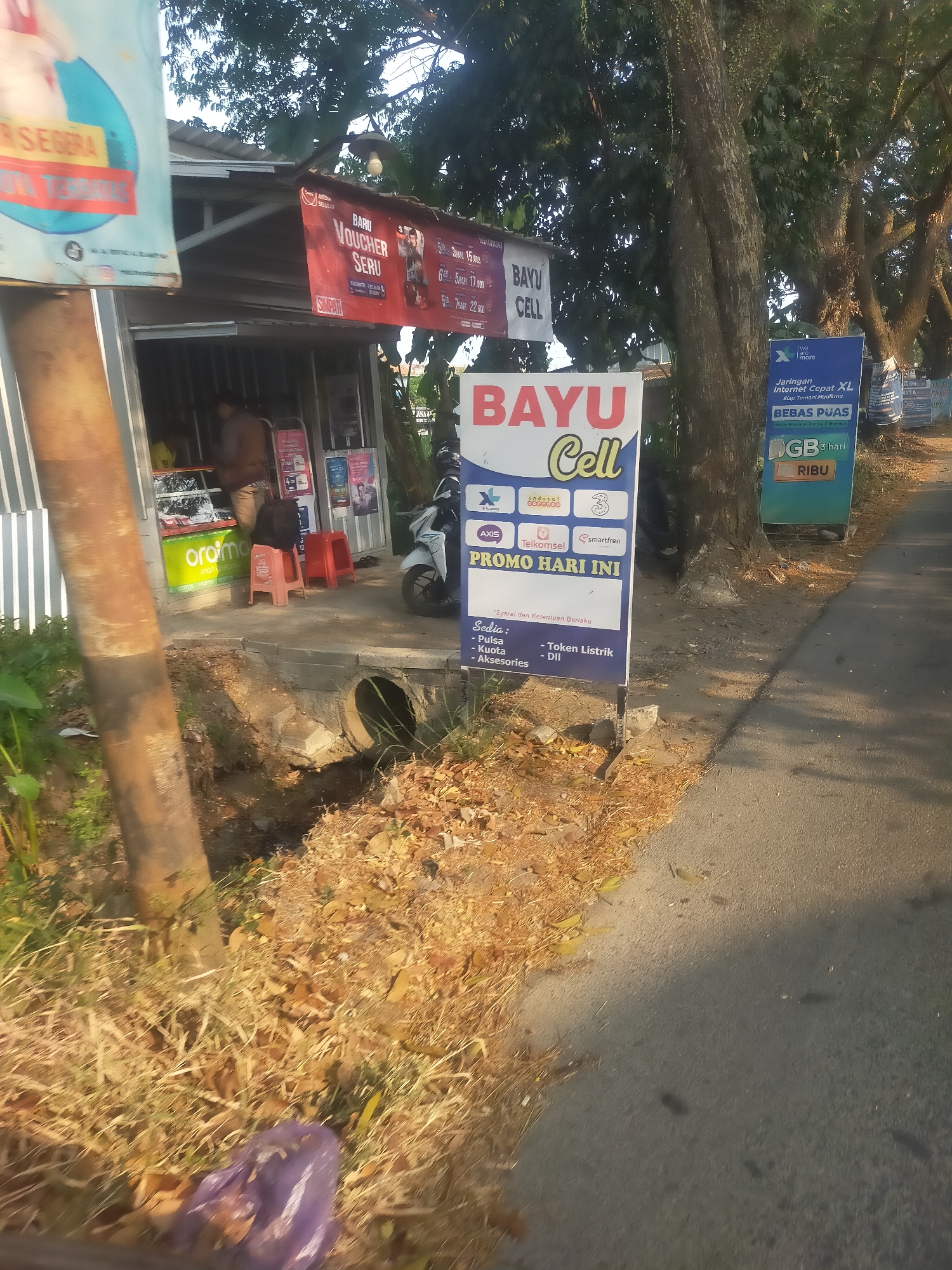 An outdoor shot captures a street scene in Kambangan, Indonesia, with a focus on a small shop and its signage. The shop appears to be a mobile phone store, as indicated by signs for "BAYU CELL" and various mobile carrier logos such as XL, Axis, Telkomsel, and Smartfren. One prominent sign advertises a "PROMO HARI INI" (Today's Promo) and lists items like "Pulsa," "Kuota," "Aksesories," and "Token Listrik" (Electricity Tokens).
The shop itself is a simple structure with an open front. Inside, a person is visible behind the counter, seemingly engaged in a transaction or arrangement. Red plastic stools are placed outside the shop, suggesting a waiting area or a place for customers to rest. A motorcycle is parked to the right of the shop.
The environment around the shop includes a paved road, a grassy area with dry leaves, and a ditch with a concrete culvert. Large trees with dense foliage line the right side of the road, casting shadows on the pavement. The lighting suggests it is daytime, possibly late morning or afternoon. The overall impression is of a typical neighborhood shop serving the local community.