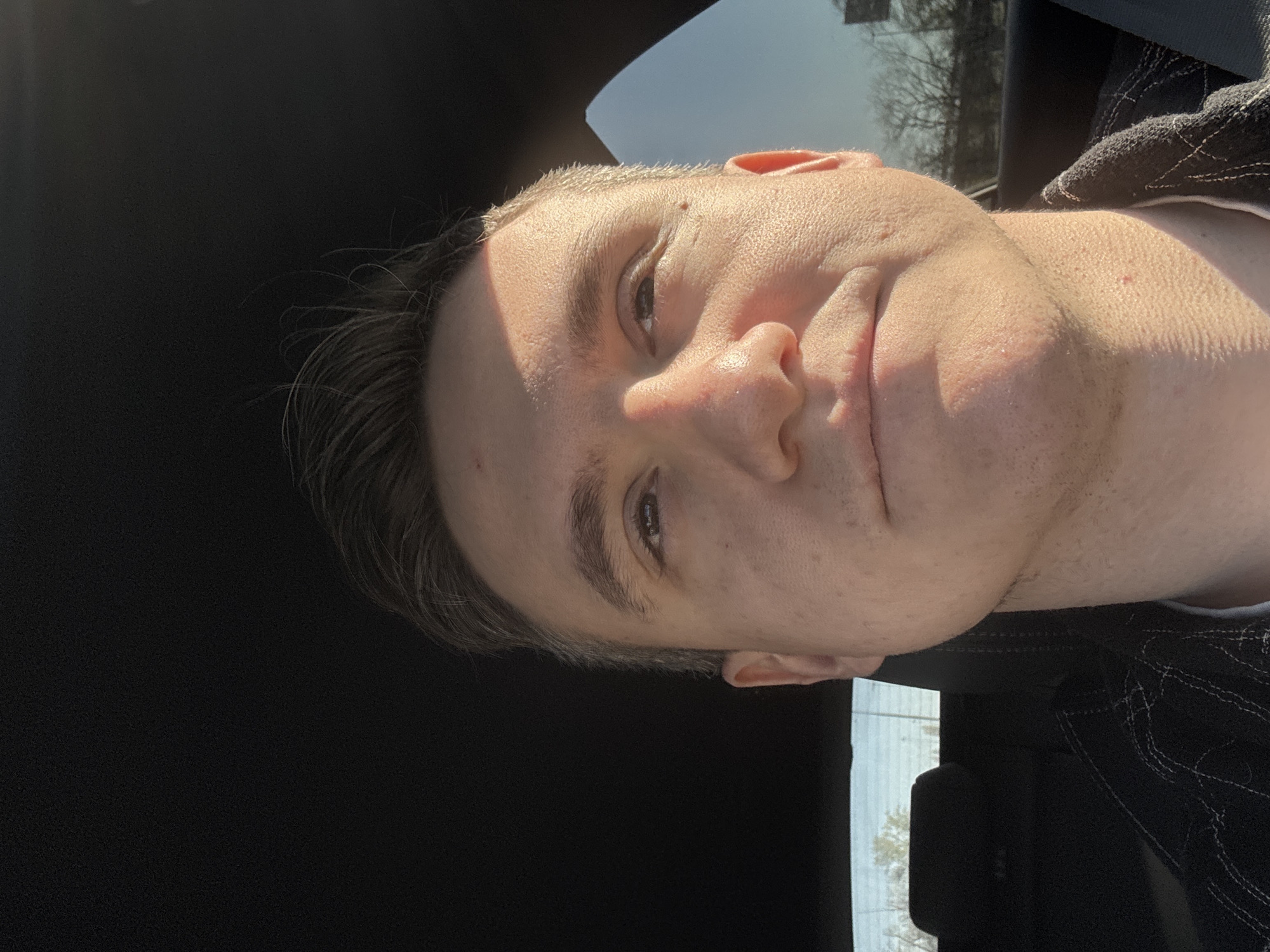 A portrait-style photograph, originally rotated 90 degrees counter-clockwise, depicts a young male seated inside a vehicle during the daytime. The man, likely in his 20s or early 30s, is positioned with his head and upper body filling the frame. He looks directly at the camera with a subtle, pleasant smile.
Strong, direct sunlight streams in from the upper right, brightly illuminating the right side of his face and shoulder, creating a stark contrast with the shadowed left side. His dark hair is styled back. He appears to be wearing a dark garment, possibly a jacket or shirt, with distinctive white stitching visible on the right shoulder area.
Through the window in the background, specifically to the left of the man, bare tree branches are visible against a bright sky, suggesting a season like late autumn, winter, or early spring. The remainder of the background consists of the dark interior of the vehicle, with parts of a car headrest or seat visible. No specific landmarks are discernible to indicate the location as Warsaw, Poland, beyond the provided context. There is no visible text.