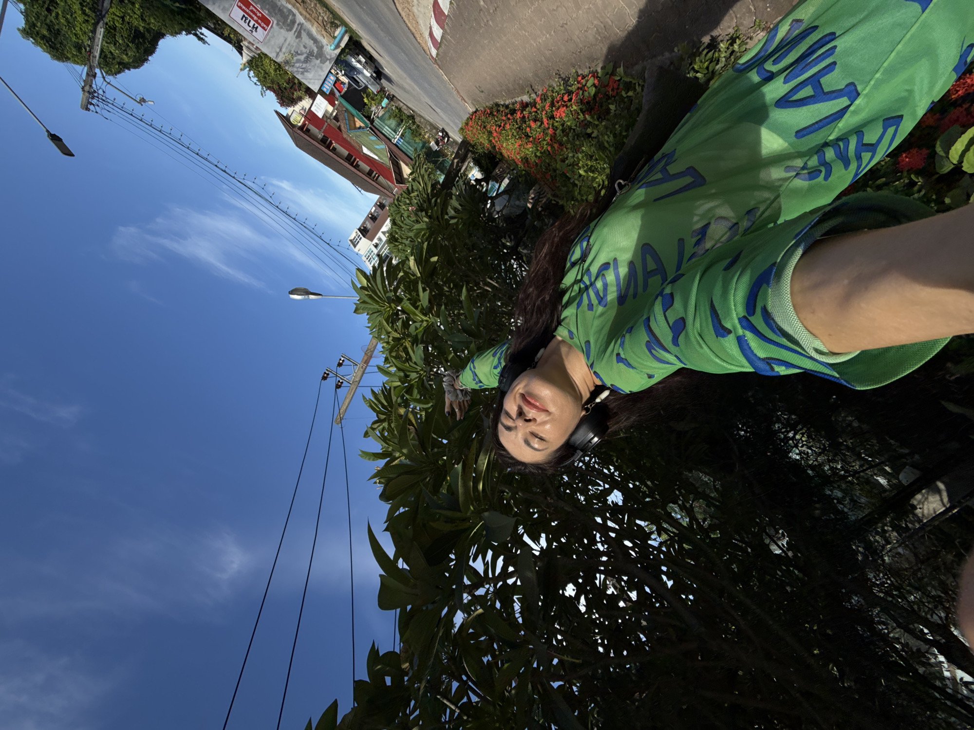 A low-angle, wide-shot selfie captures a woman in Karon, Thailand, standing outdoors near a lush green tree and a flowerbed. The woman, wearing black headphones and a sheer green mesh shirt with blue lettering, is looking up at the bright blue sky with her eyes closed and a slight smile. Her right arm is raised, with her hand extended towards the sky and her fingers splayed. Behind her, a street is visible with a sidewalk, a road, buildings, and utility poles with wires. The sun is casting strong shadows, indicating it is daytime.