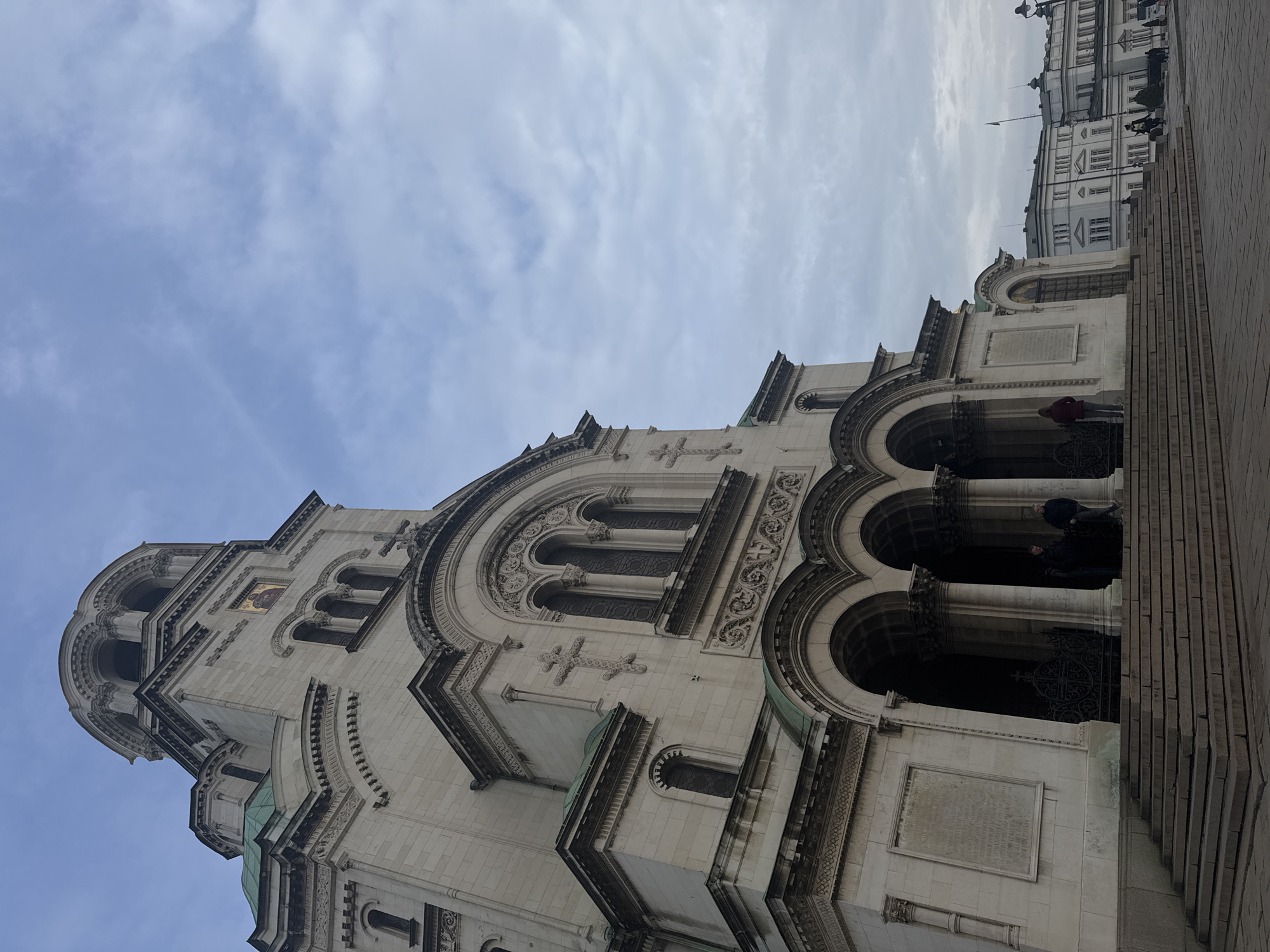 The image shows a low-angle view of the Alexander Nevsky Cathedral in Sofia, Bulgaria. The cathedral's ornate facade, with arches, crosses, and decorative elements, is prominently featured. People can be seen on the steps and near the entrance of the cathedral. The sky is overcast. Adjacent buildings are visible in the background.
