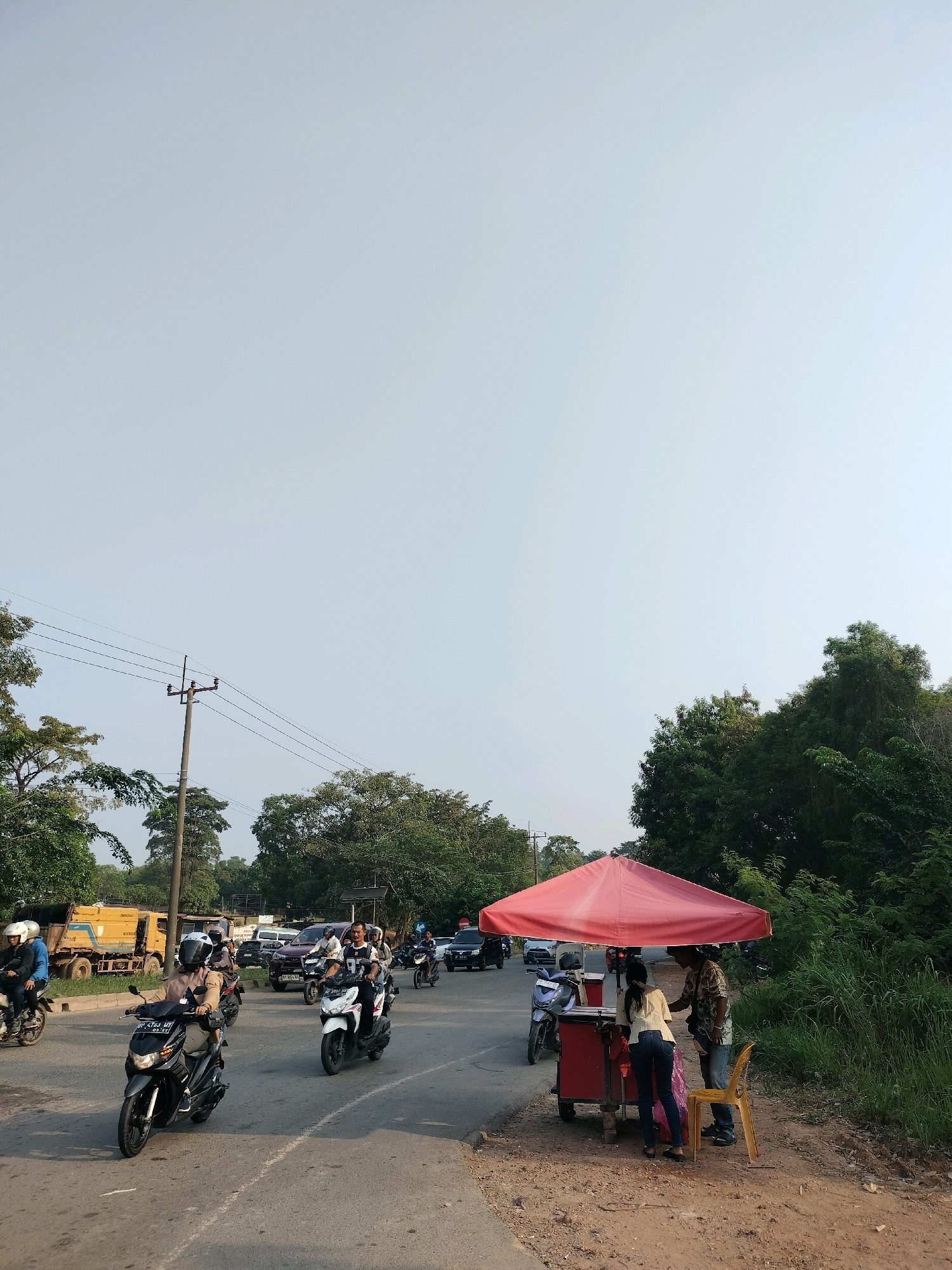 The image captures a street scene in Batam, Indonesia. A red umbrella covers a food cart on the side of a road, where two individuals are interacting, possibly a vendor and a customer. A yellow chair is placed next to the cart. The road is populated with several motorcycles, with some riders wearing helmets. In the background, there is traffic including cars and a yellow truck. Lush green trees and vegetation line the roadside, and a utility pole with wires is visible. The sky is clear and bright.