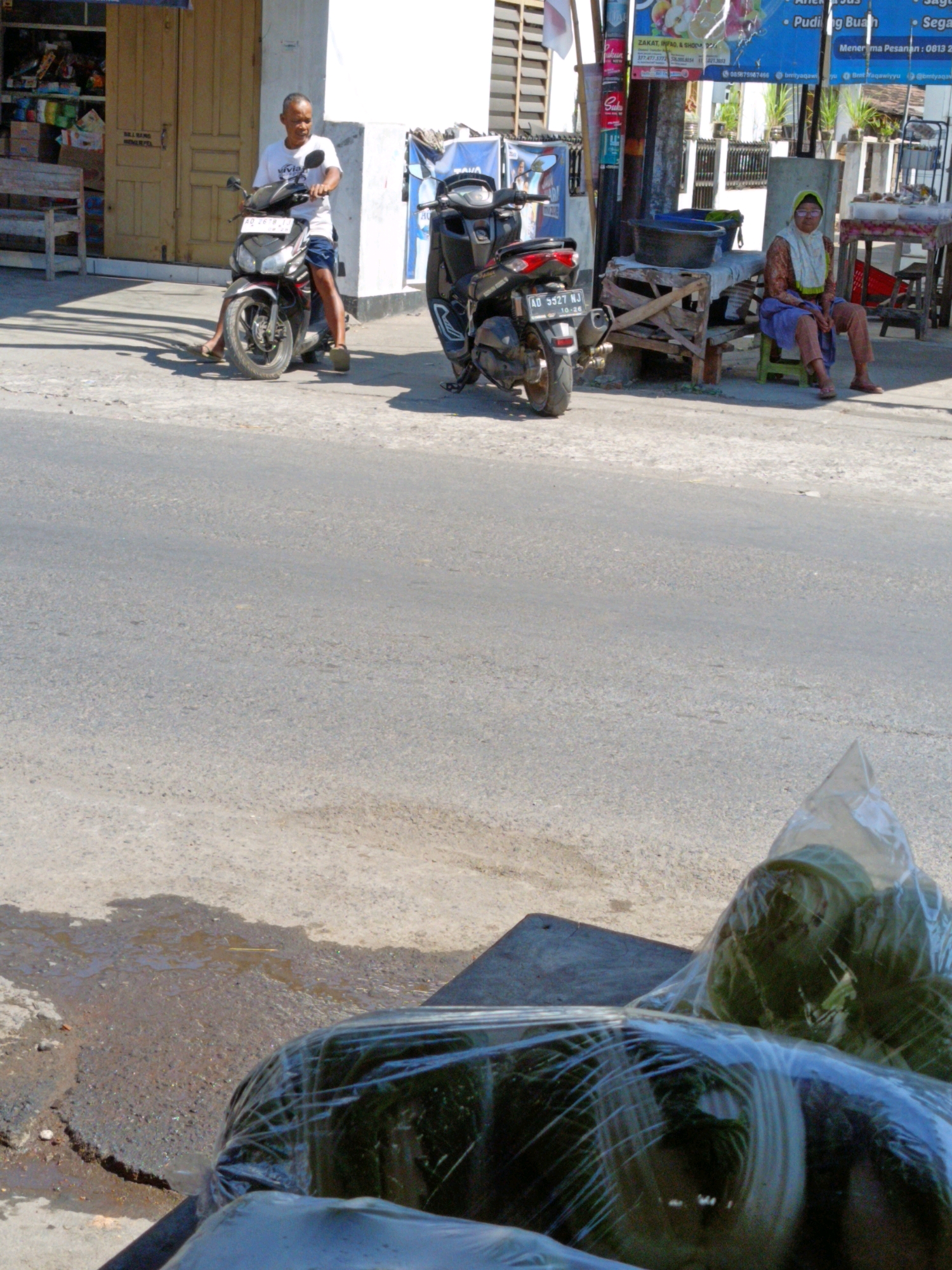 In Sukorini, Indonesia, a man rides a black motorcycle on a street. He wears a white t-shirt and blue shorts. To his right, a woman in a headscarf and patterned clothing sits on a green stool. Behind her, produce is displayed on a wooden stand. In the foreground, a black plastic bag filled with green vegetables rests on the ground. The street is paved and shows signs of wear. The background features buildings and street signs.