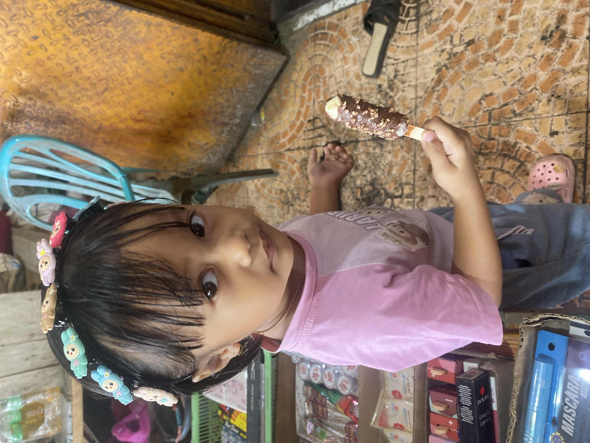 In Ta', Indonesia, a young girl with dark hair adorned with colorful animal-shaped hair clips stands in what appears to be a market or shop. She is wearing a pink t-shirt and grey pants, holding a chocolate-covered popsicle with nuts. The background reveals shelves stocked with various products, including makeup and small containers. A turquoise plastic chair is visible behind her, next to a textured, yellow wall. The floor is tiled with a patterned design in shades of orange and brown. A black sandal lies on the floor to the right of the girl.