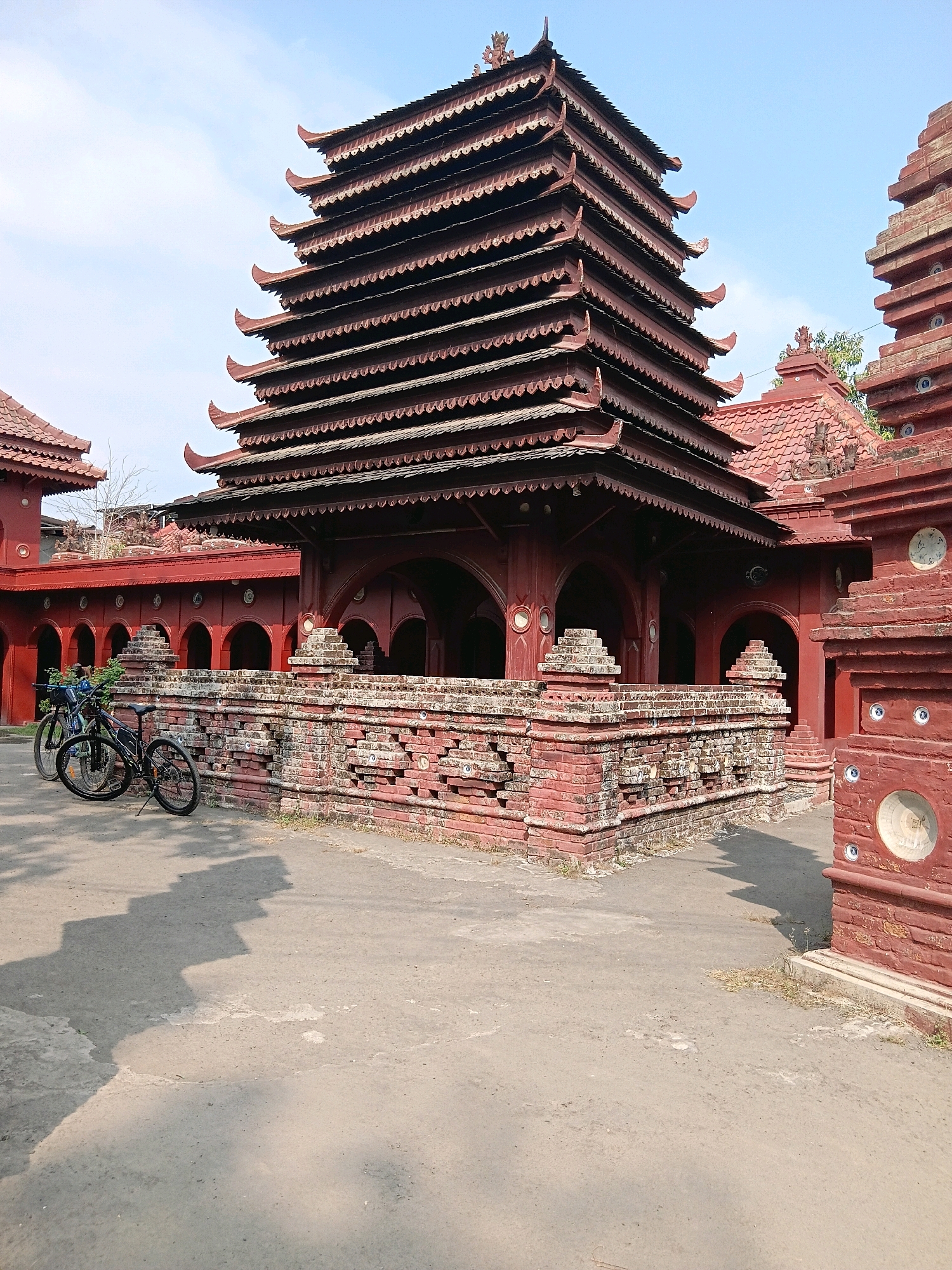 The image shows the exterior of the Sunan Gunung Jati Mausoleum complex in Cirebon, Indonesia. A multi-tiered, dark red pagoda-style building dominates the center of the frame, with intricately carved roof edges. To the left and right of this central structure are other red brick buildings with tiled roofs. A low, decorative brick wall with layered horizontal elements runs in front of the central building. Two bicycles are parked beside this wall. The ground is paved with concrete. The sky is clear and blue. The overall impression is of a well-maintained historical and religious site under a bright sky.