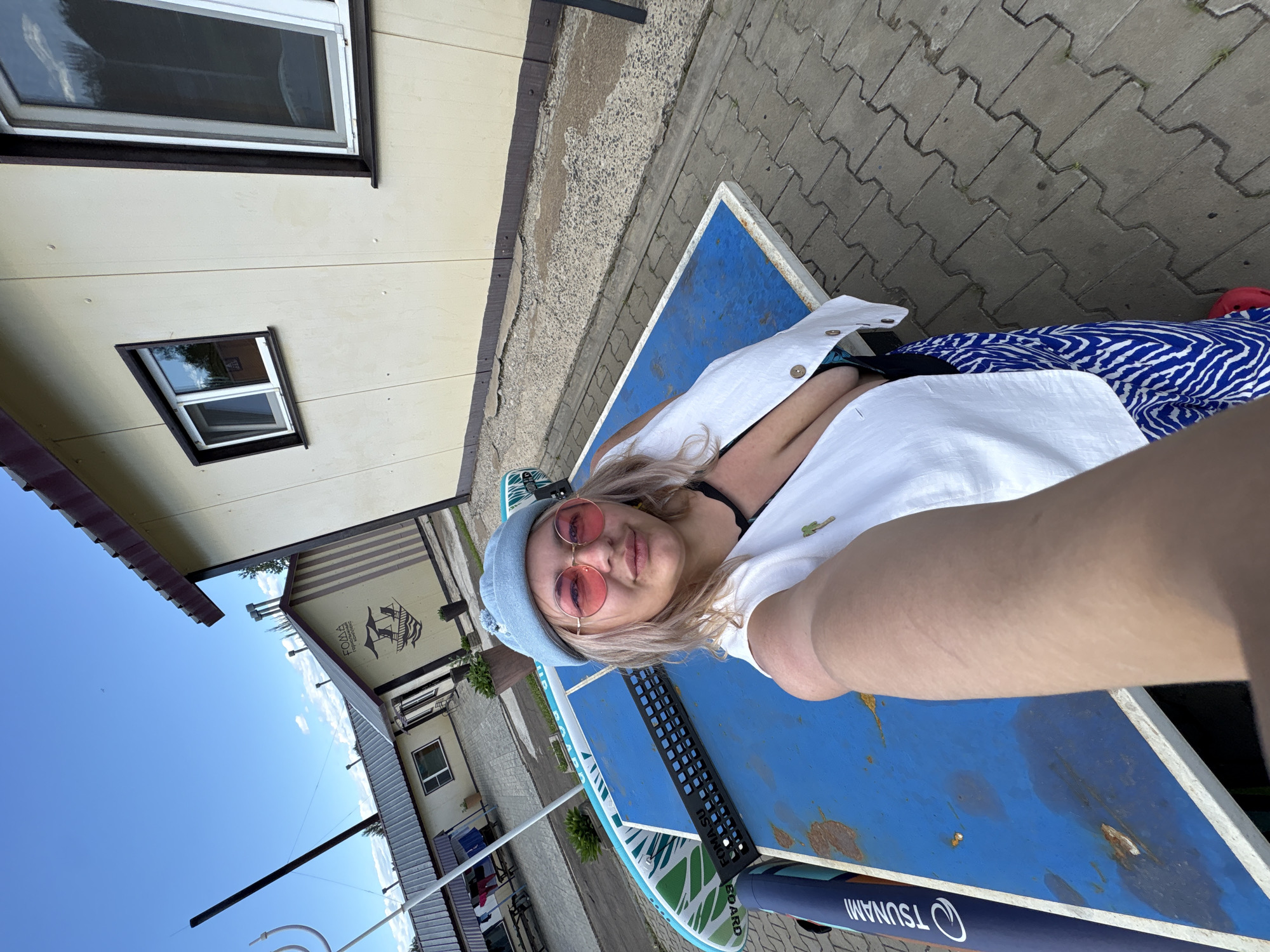 A woman in a blue hat and pink sunglasses takes a selfie in front of a ping pong table with a paddleboard attached. She wears a white open vest over a dark top and a blue and white patterned skirt. The background shows a light-colored building with windows and a clear sky with scattered clouds. The scene appears to be outdoors, possibly at a recreational facility.