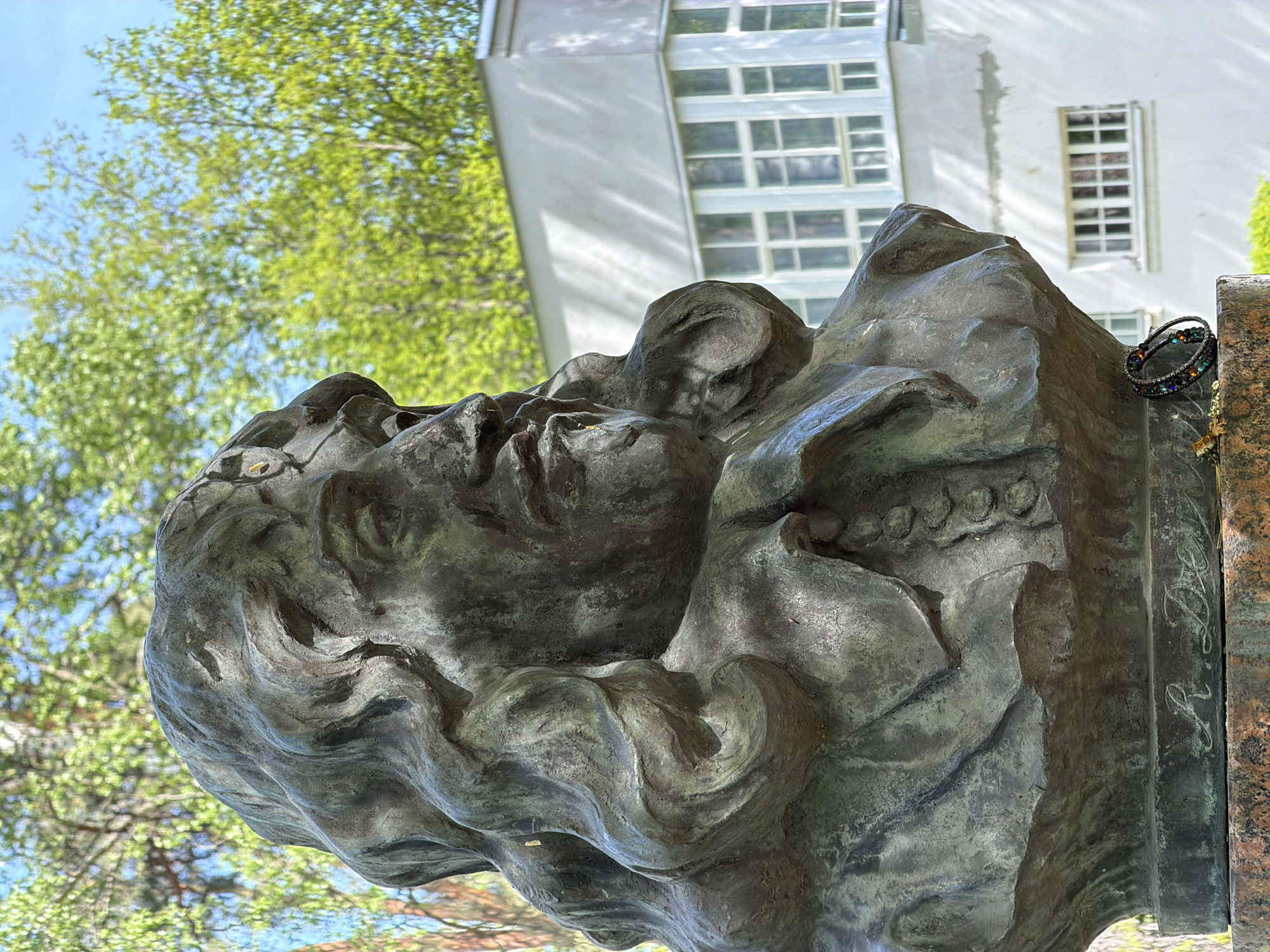 The image depicts a bronze sculpture of a human face, possibly a monument or memorial. It appears to be a depiction of a man with a weary expression. The sculpture is set against a backdrop of green foliage and a white building with a grid-like window pattern. A bracelet is resting on the edge of the bronze sculpture. The photograph appears to be taken from a low angle, looking up at the sculpture. The sculpture is the monument to Anton Chekhov in Tokkari, Russia.