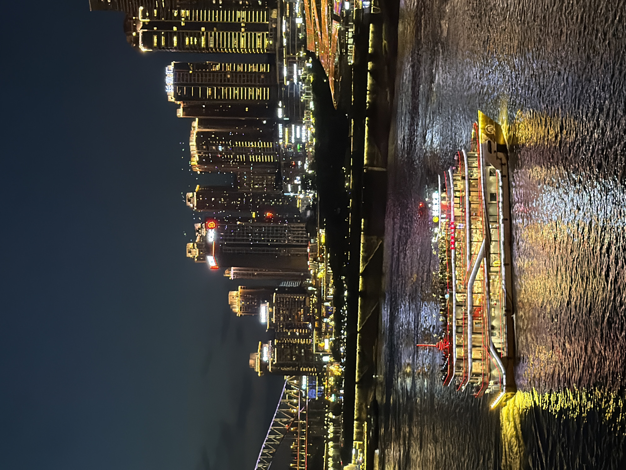 The image depicts a nighttime scene along the Yangtze River in Chongqing's Yuzhong District, China. A multi-decked tour boat, illuminated with numerous yellow and red lights, cruises across the dark water, leaving a trail of yellow light in its wake. The boat is filled with visible passengers on its upper decks. In the background, the illuminated skyline of Chongqing rises, featuring a dense collection of modern skyscrapers adorned with bright lights. A large bridge with illuminated arches spans the river in the mid-ground, connecting the city. The water reflects the vibrant lights from the city and the boat, creating a shimmering effect on the surface. The overall atmosphere is one of a bustling urban nightscape.