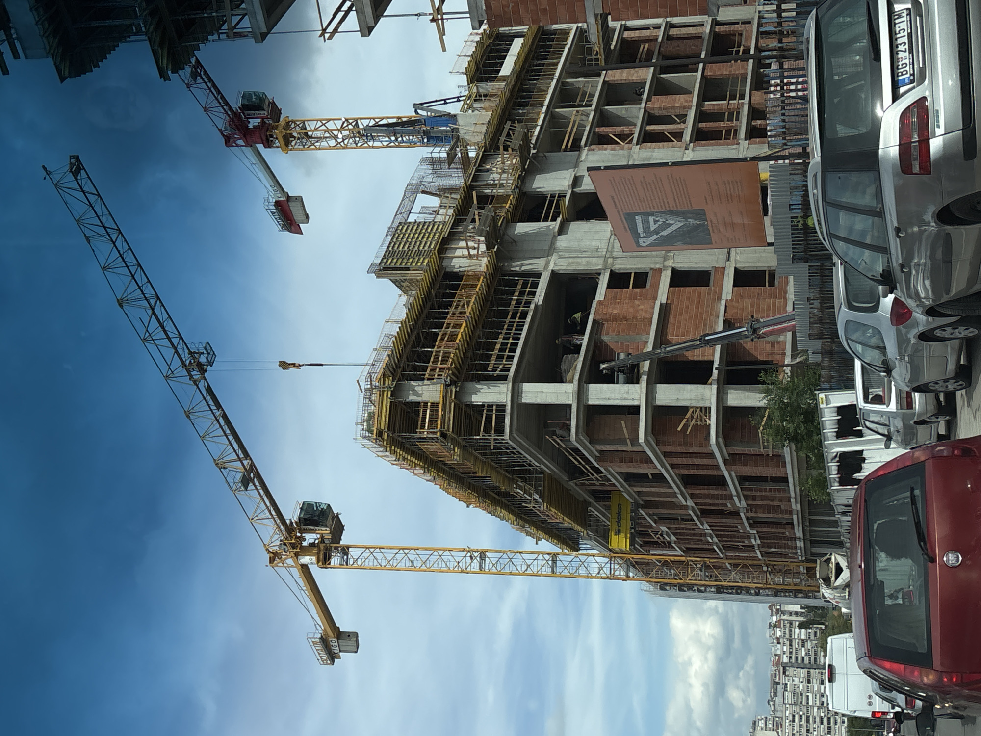 The image shows a construction site in Vozdovac Urban Municipality, Serbia. A multi-story building is under construction, with visible brickwork and concrete structures. Several tower cranes are present, including one large yellow crane dominating the left side of the image. The sky is partly cloudy. Cars are parked on a street adjacent to the construction site, and a partially obscured cityscape is visible in the background.