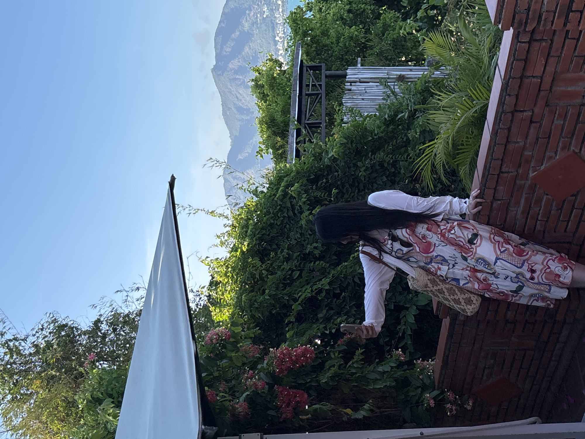A woman with long dark hair stands on a brick structure beside a dense green foliage. She wears a white long-sleeved shirt and a patterned dress. She holds a smartphone in her right hand. A mountain is visible in the background under a clear sky. The scene appears to be set near Bac Nha Trang Ward, Vietnam, based on the environmental elements.