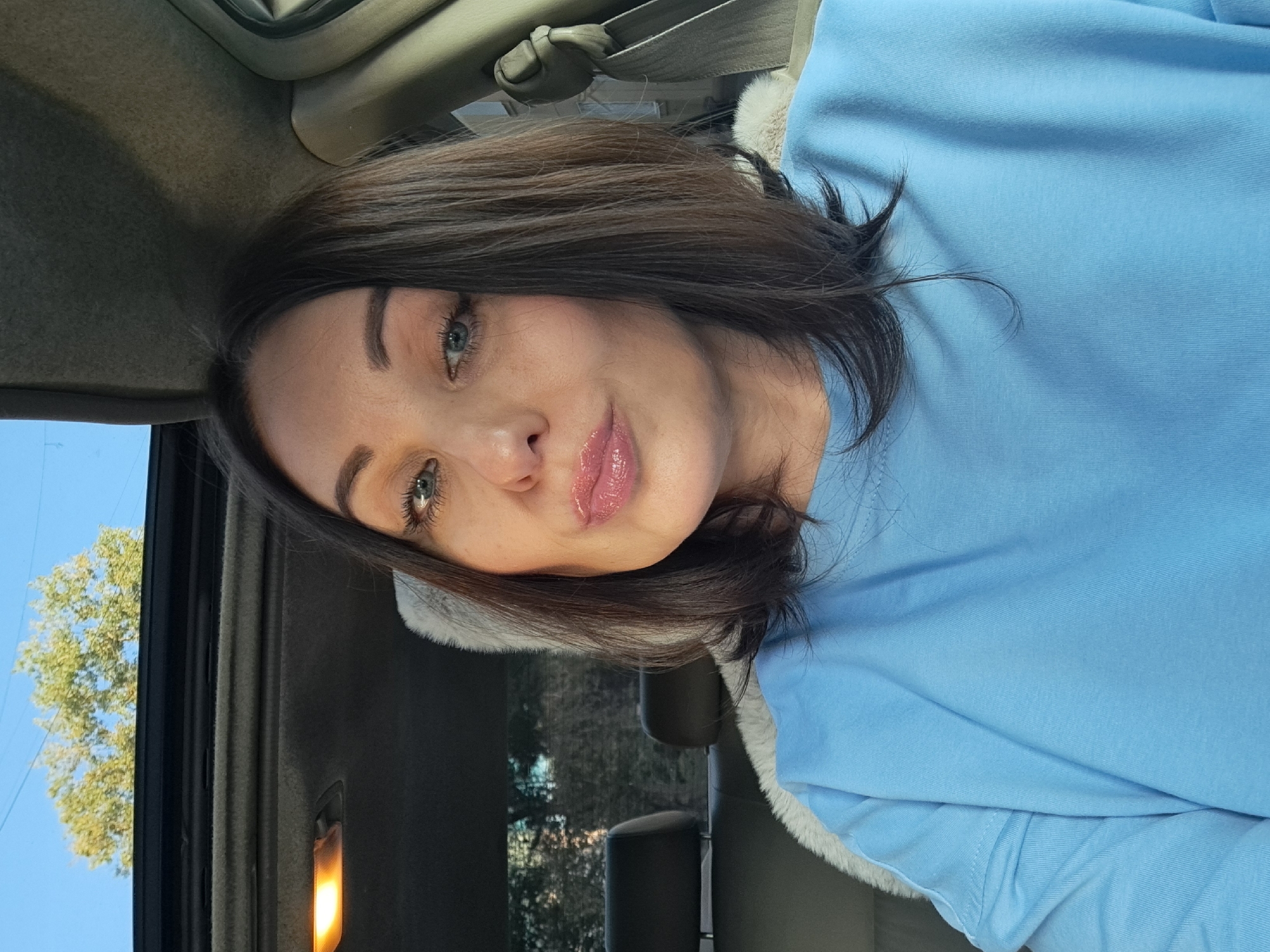 A woman with shoulder-length dark hair and blue eyes, wearing a light blue sweatshirt, takes a selfie inside a car. The car's sunroof is open, revealing a clear blue sky and the top of a leafy green tree. Inside the car, the ceiling panel and a dome light are visible. The woman is sitting in the driver's seat, and the passenger seat headrest is visible behind her, covered with a fluffy white material. The interior of the car, including a seatbelt, is also visible. The background outside the car shows trees and foliage, suggesting an outdoor location, possibly in the city of Irkutsk, Russia, given the context of the prompt.
