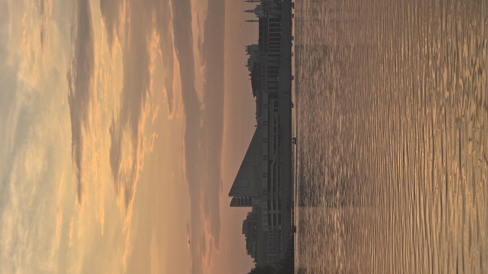 The image depicts a wide-angle view of the Kazanka River in Kazan, Russia, during a sunset or sunrise. The sky is filled with soft, peachy-orange clouds, indicating a warm, diffused light. In the middle ground, a long, modern building with a distinctive angled roofline dominates the skyline, likely the Kazan Family Center. To the right of this structure, a series of older buildings and minarets of a mosque are visible, suggesting a mix of modern and historical architecture along the waterfront. The river itself, in the foreground, is calm and reflects the warm colors of the sky, creating a rippling, textured surface. Several small boats are scattered across the water, with a few tiny figures visible within them, suggesting leisure activities. The overall scene is serene and picturesque, capturing the ambiance of the city at twilight.