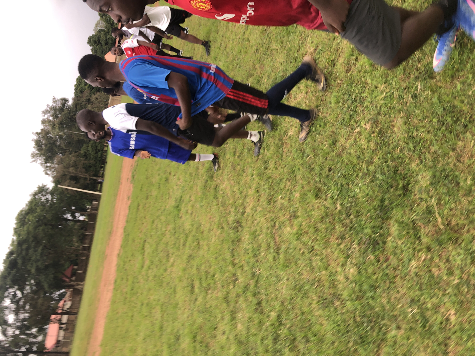 In Jos, Nigeria, a group of young men are engaged in a soccer practice on a grassy field adjacent to a dirt track. The individuals, dressed in soccer attire including jerseys and shorts, are captured in motion, suggesting an active training session. The environment is characterized by lush green grass and trees in the background, with buildings visible in the distance, indicative of a semi-urban or suburban setting. The lighting suggests an overcast day.