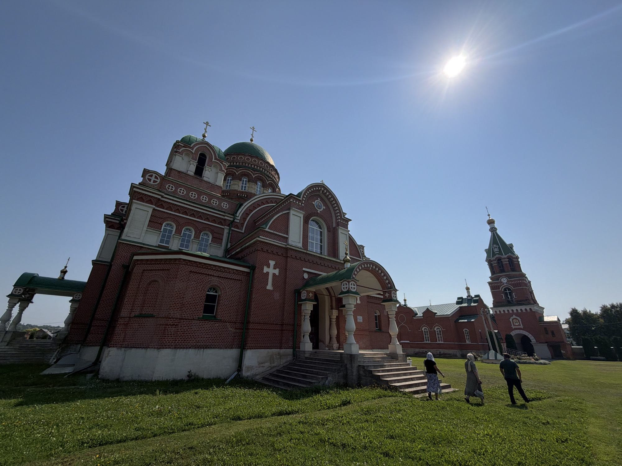 The image depicts a wide-angle view of the Resurrection Convent in Troekurovo, Russia, on a clear, sunny day. The main subject is the large, red brick cathedral with green domes, characteristic of Russian Orthodox architecture. To the right, a tall, red brick bell tower with a green spire stands prominently. Several individuals are present in the foreground, walking on the grassy grounds. The environment is characterized by lush green grass, a bright blue sky, and the imposing religious structures. The sun is visible in the upper right corner, casting a bright glare. The overall scene conveys a sense of tranquility and religious significance.