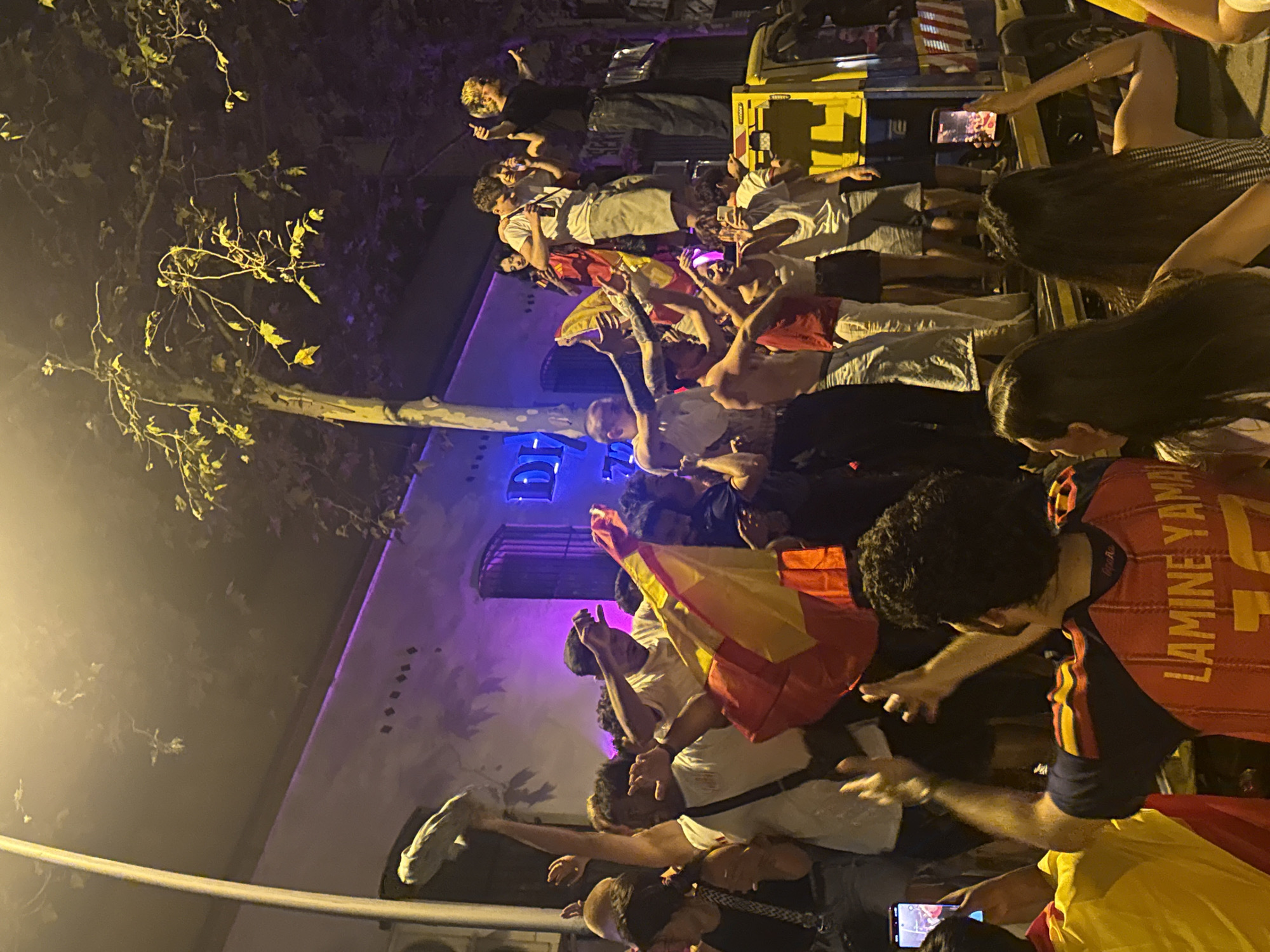 The image captures a lively street scene at night in Barcelona, Spain, with a crowd of people gathered around a yellow truck. Many individuals are holding Spanish flags, waving them enthusiastically, and appear to be celebrating. Some are on top of the truck, while others are on the street level. The atmosphere is energetic, with people cheering and taking pictures with their phones. The background shows a building with purple lighting and trees with illuminated leaves, suggesting a festive or event-related gathering. The overall impression is one of collective joy and celebration in a public urban space.