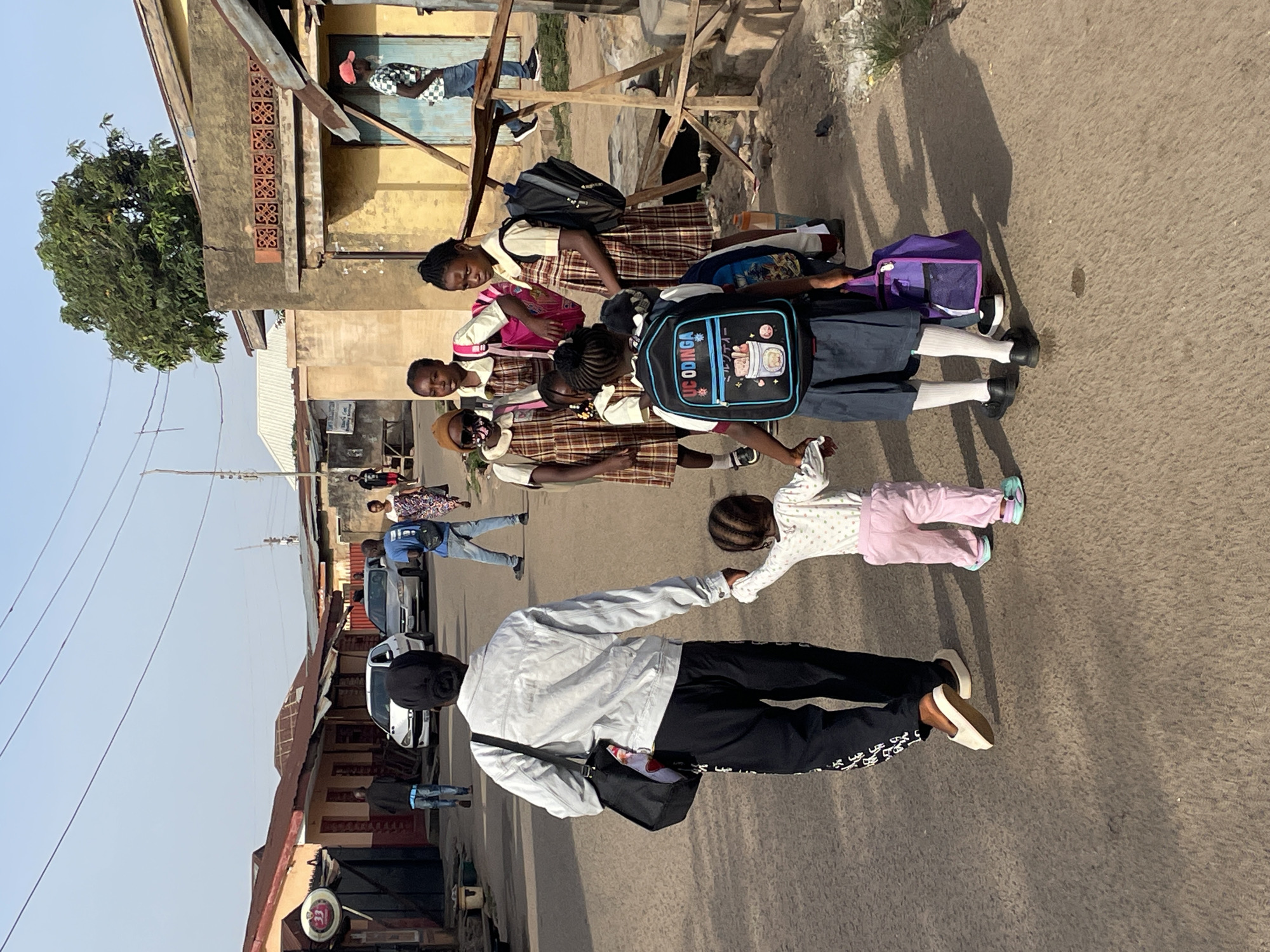 This image captures a vibrant street scene in Jos, Nigeria, likely during school hours, under bright daylight. The main subjects are a group of several schoolgirls, accompanied by adults and a small child, walking along a paved road.
The schoolgirls are dressed in similar uniforms, consisting of plaid brown, white, and red dresses or skirts paired with white shirts. Some wear dark blue or black skirts/trousers as part of their uniform. They carry various backpacks and bags; one black backpack prominently displays the word "ODINGA" in white, along with Japanese text and a cartoon character design. Another girl carries a purple sports-style bag.
In the immediate foreground, an adult wearing a light-colored jacket, black pants with white patterns, and a black cap walks while holding the hand of a very young child. The child is dressed in a light-colored top with pink bottoms and light blue shoes. Their shadows are visible on the ground, indicating a sunny day.
The setting is an urban or semi-urban street, with various buildings lining the road. These structures appear weathered, some with exposed brickwork or concrete and corrugated metal roofs. Power lines are visible stretching across the clear, bright sky. Several cars are parked along the street, and other pedestrians, including a man in a blue shirt and jeans, and another in a checkered shirt in a doorway, can be seen in the background. A large green tree stands tall on the left side of the frame. The overall atmosphere is calm and depicts typical daily activities.