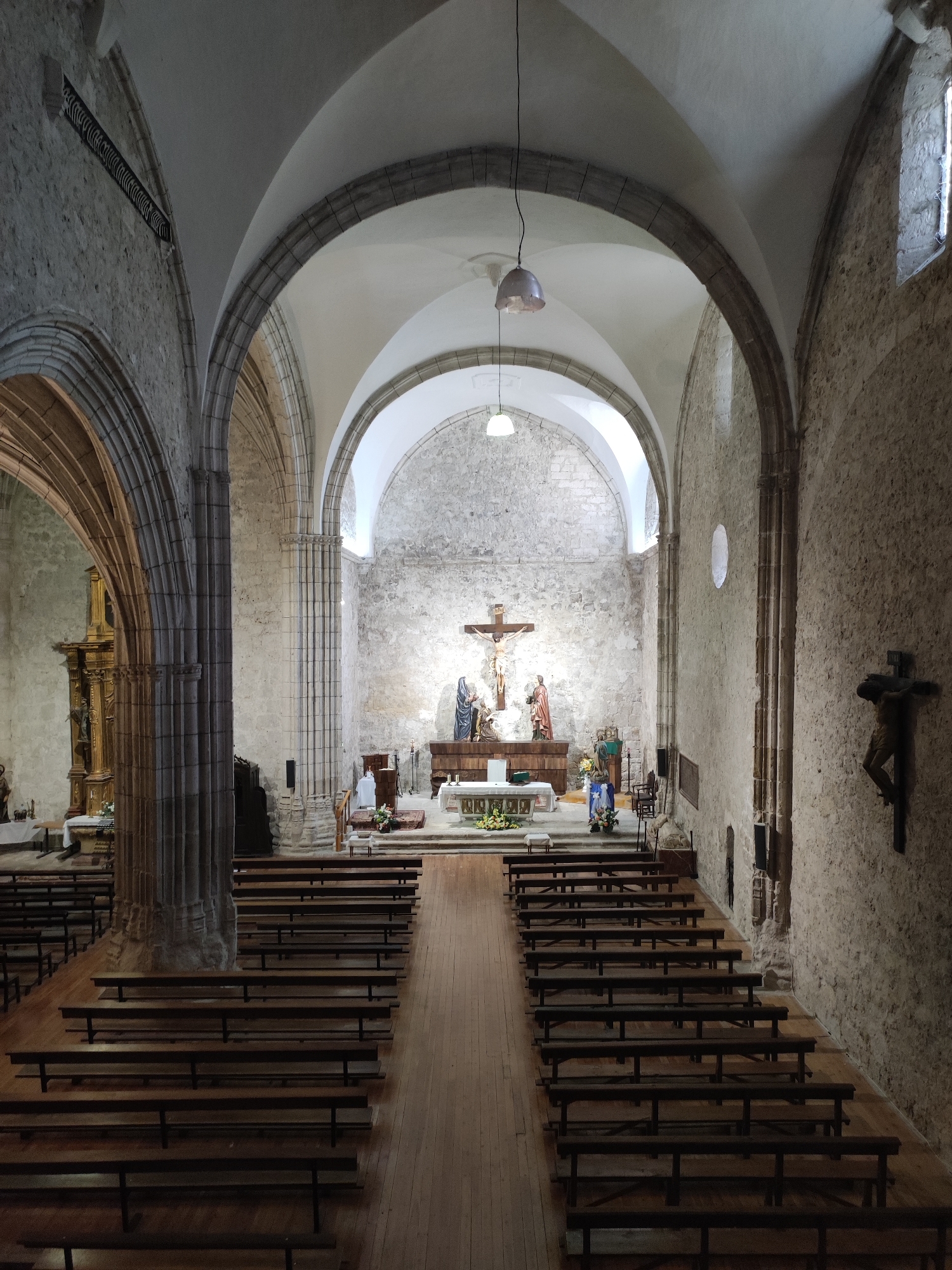 This image shows the interior of the Church of Nuestra Señora de la Asunción in Traspinedo, Spain. The church features a nave with stone walls and arched ceilings. Rows of wooden pews fill the nave. At the far end, a stone altar is visible, with a crucifix and statues of religious figures. A smaller crucifix hangs on the right wall. Natural light enters through a window on the right wall. The floor is made of wooden planks.