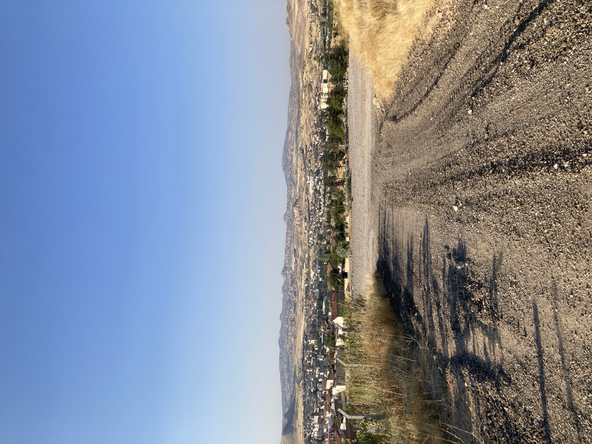 The image captures a wide-angle view of a landscape with a prominent dirt road in the foreground. The road, composed of gravel and dirt, exhibits tire tracks and is illuminated by sunlight, casting shadows across its surface. Dry, golden grass lines the right side of the road, while similar vegetation, interspersed with a wire fence, occupies the left foreground.
In the middle ground, a town is visible, characterized by numerous buildings with red-tiled roofs, indicative of typical Turkish architecture. The town stretches across a valley, bordered by hills in the distance. These hills appear arid and rocky, with distinct geological formations that are characteristic of the Cappadocia region. A highway is discernible, cutting through the town and leading into the distance.
The background is dominated by a clear, bright blue sky, suggesting fair weather and good visibility. The overall scene depicts a rural or semi-rural setting in Avanos İlçe Merkezi, Turkey, highlighting the contrast between the man-made structures of the town and the natural, rugged terrain of the surrounding landscape. There are no people or animals visible in the image.