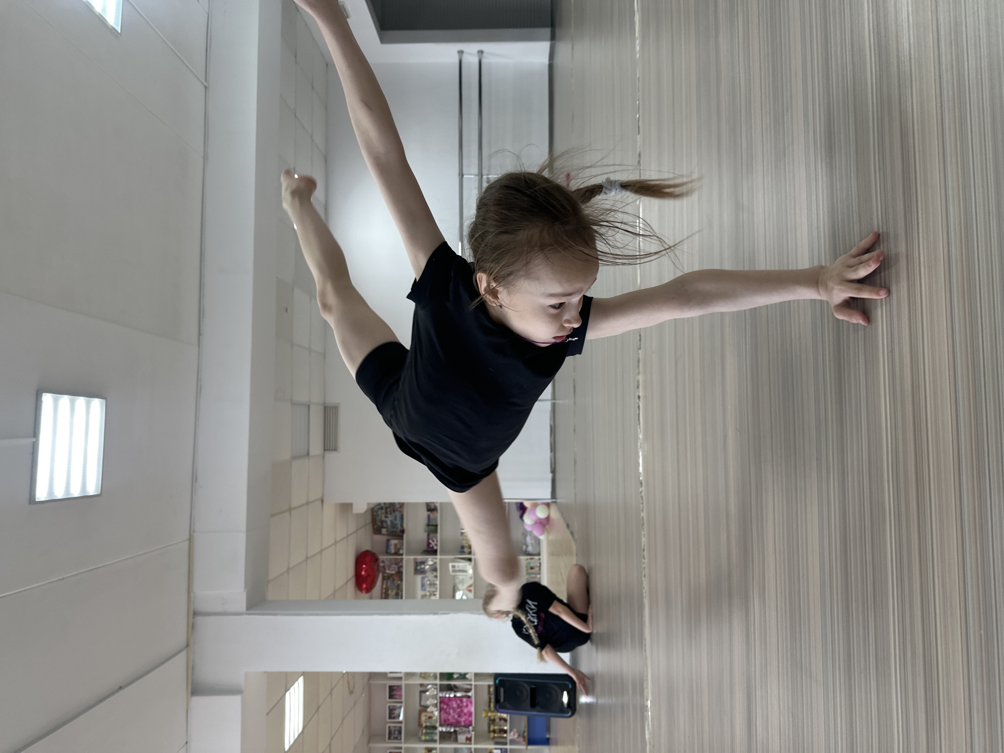 A young girl in a black outfit is performing a split against a light-colored wall in a room with a light-colored wood floor. Her right hand is touching the wall. Behind her and to the left, another person is kneeling on the floor. There is a boombox nearby. Shelves filled with items are visible in the background. Based on the items visible in the background, the location is likely a dance studio in Votkinsk, Russia.