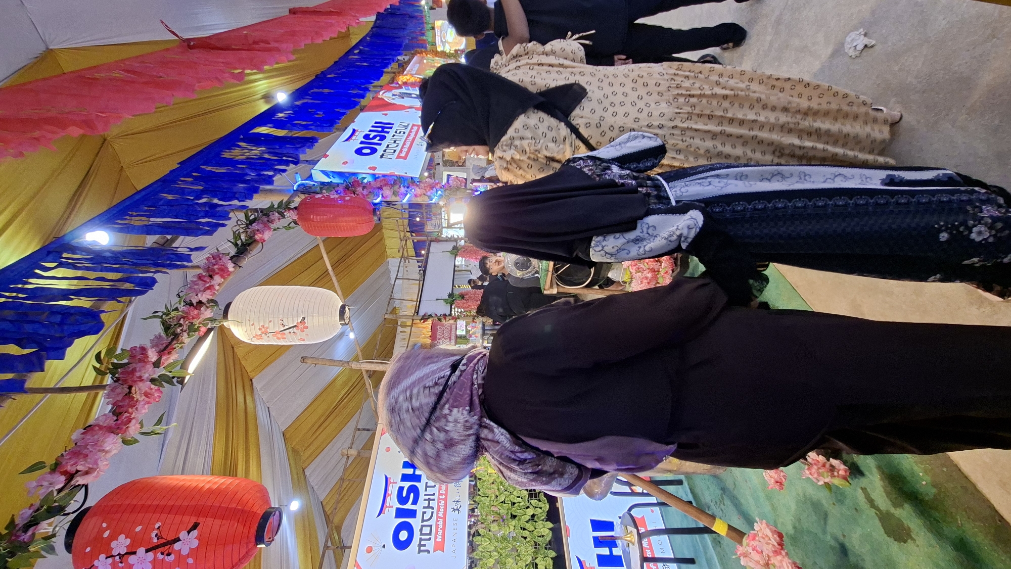 This is a medium shot, eye-level, outdoor photograph taken at a street market in Lhokseumawe, Indonesia. The image captures a scene with several people walking through a decorated stall area. The overhead view of the market is visible, with yellow and white striped fabric forming a canopy, adorned with blue and pink paper streamers and artificial cherry blossoms. Red and white paper lanterns with floral designs hang from the ceiling, adding to the festive atmosphere.
In the foreground, three individuals are the primary subjects. On the left, a person wearing a dark purple hijab and a black abaya is seen from behind, with their left arm extended. To their right, a person in a black hijab and a black and white patterned dress is walking, holding hands with the person to their right. The third person, also seen from behind, wears a beige patterned dress and a black hijab. Their right arm is around the shoulder of the person in the patterned dress.
In the midground, other people are visible, including a man in a black shirt looking towards the camera. Behind him, there is a stall with a sign that reads "OISHI MOCHISUKI". The stall features greenery, likely plants, and a display of goods. Various other stalls and people can be seen further in the background, suggesting a busy market environment. The ground is a textured concrete or asphalt surface. The overall lighting suggests it is daytime, with artificial lights from the market adding to the ambiance.