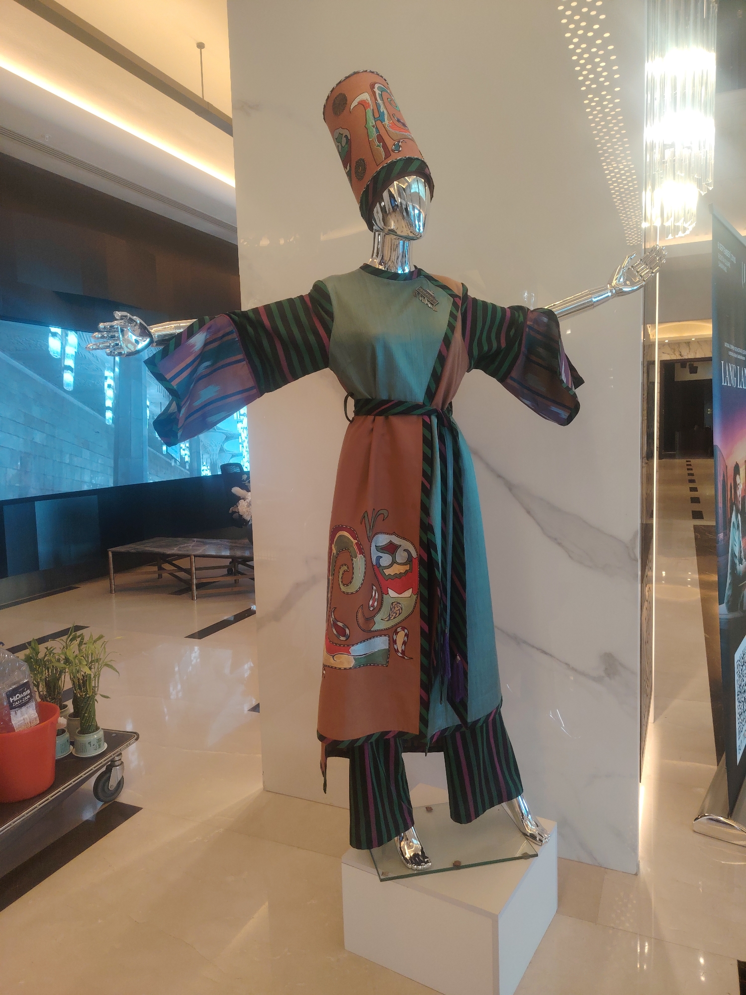 This image shows a mannequin dressed in a vibrantly colored, traditional-style outfit, likely displayed in a public space such as a hotel lobby or a retail store. The mannequin is metallic and features articulated limbs. It is wearing a tall, decorative hat with an orange base and colorful embroidered motifs. The clothing consists of a teal tunic over striped trousers, with a brown overdress featuring a bold paisley-like print. A sash tied around the waist completes the ensemble.
The background includes a modern interior with polished floors, marble-patterned walls, and bright, contemporary lighting fixtures, including a striking crystal chandelier. In the lower left corner, a red bucket holds plants and a bottle of water. To the right, a poster with text and an image is partially visible. The lighting suggests an indoor setting, possibly during the day, with artificial light contributing significantly to the scene's illumination.
There are no people visible in the image; the central subject is the mannequin. The overall impression is a carefully curated display designed to showcase the ethnic attire.
The prompt requests information about the location in the city of Konigil, Uzbekistan. However, the image itself provides no direct visual cues or contextual information to place it specifically in Konigil. The interior design and the style of the displayed clothing might suggest Central Asian influence, but without additional metadata or explicit signage within the image, pinpointing the exact city and country is not possible based solely on the visual evidence.