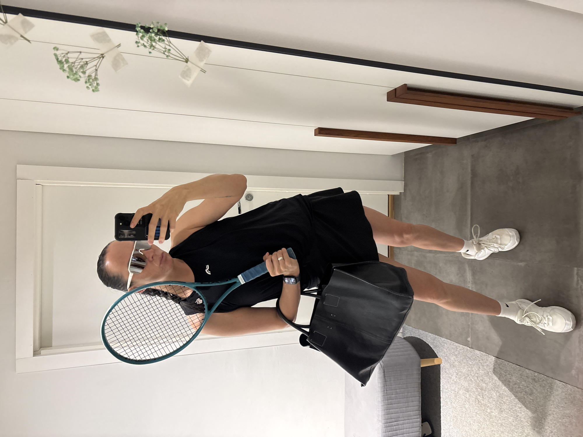 A person is taking a selfie in a mirror. The person is wearing white-framed sunglasses, a black tennis dress, and white sneakers. They are holding a tennis racket and a black leather tote bag. The background includes a white wall, a gray floor, and a grey upholstered bench. Baby's breath is taped to the mirror. The location is unknown and cannot be determined from the image.