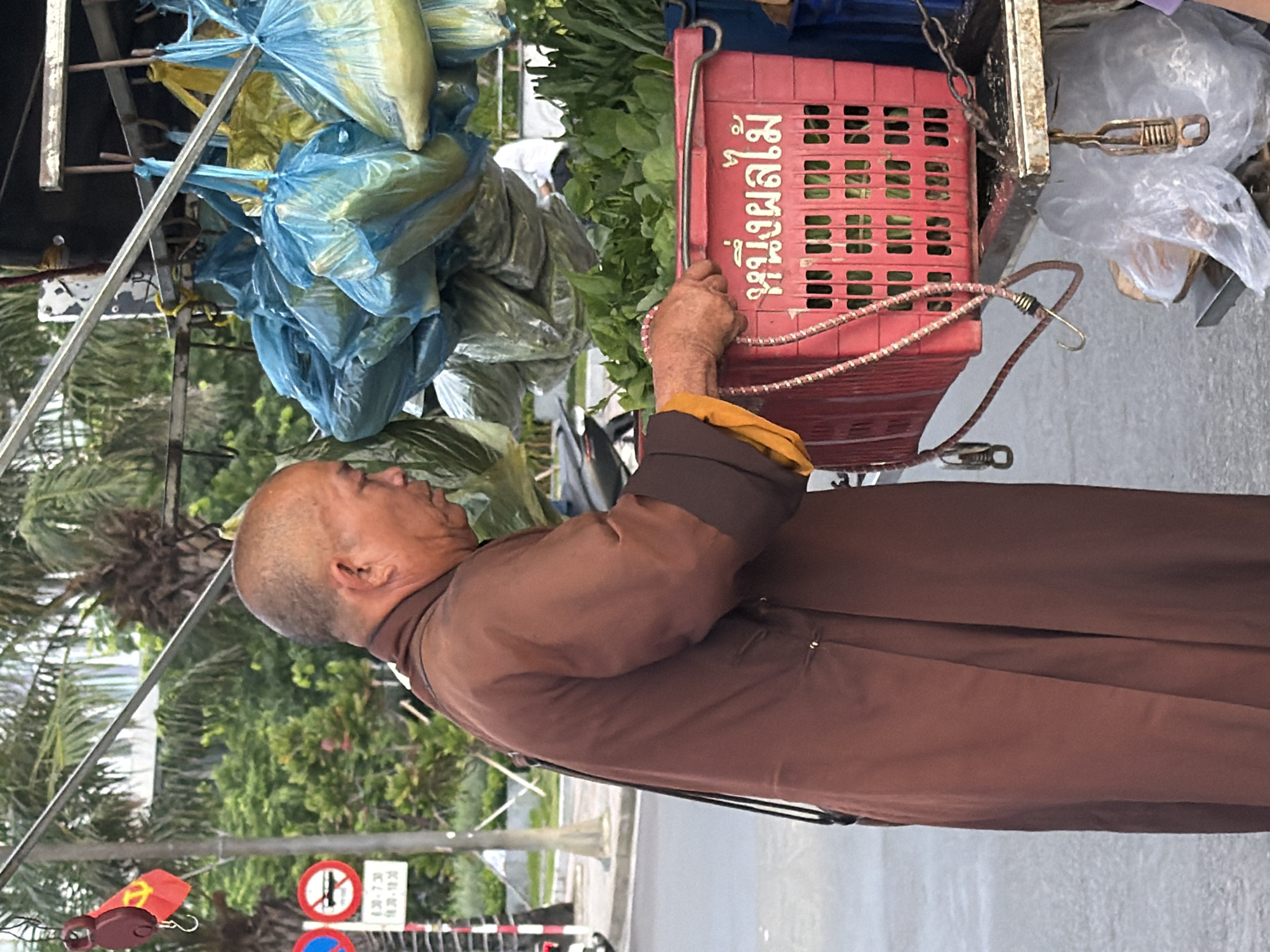 A bald man wearing a brown robe and a yellow sash is seen from the side, reaching into a red plastic basket filled with green vegetables. The basket has white Thai writing on it. The man is standing on a street in Nha Trang, Vietnam, next to a market stall. The stall has blue and yellow plastic bags filled with produce hanging from a metal frame. In the background, there are palm trees and other greenery, suggesting a tropical climate. A traffic sign with red and white stripes is visible to the left of the man. The overall scene depicts a street vendor selling fresh produce in a bustling urban environment.