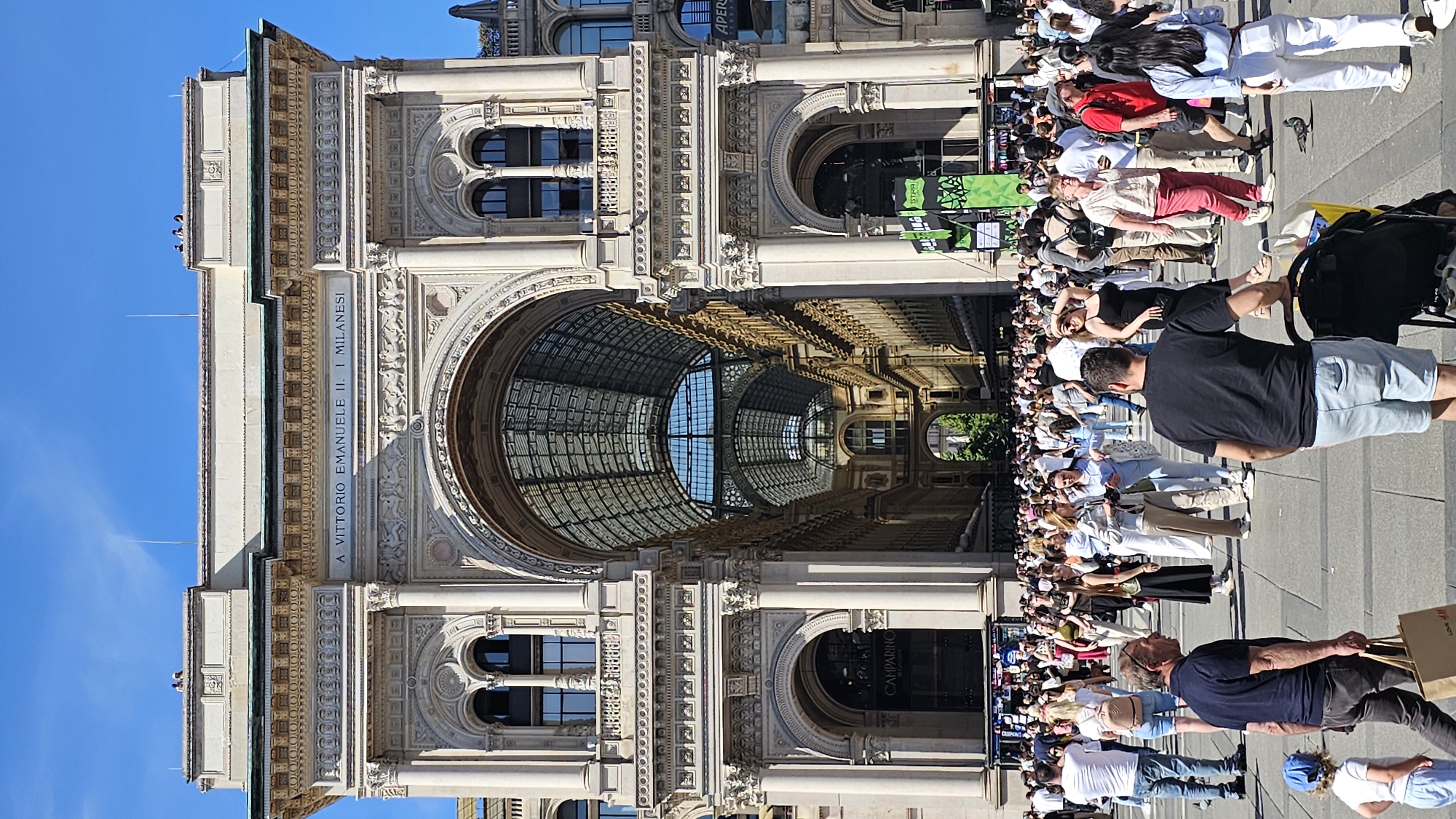 The image captures a bustling scene in the Galleria Vittorio Emanuele II in Milan, Italy. The Galleria's ornate architecture is visible, including the glass dome and detailed facades. A crowd of people is present in the foreground, some walking, some standing, and others sitting, with varying clothing and personal belongings. The clear sky suggests the photo was taken on a bright day. The scene appears to be a typical day at this popular tourist destination.